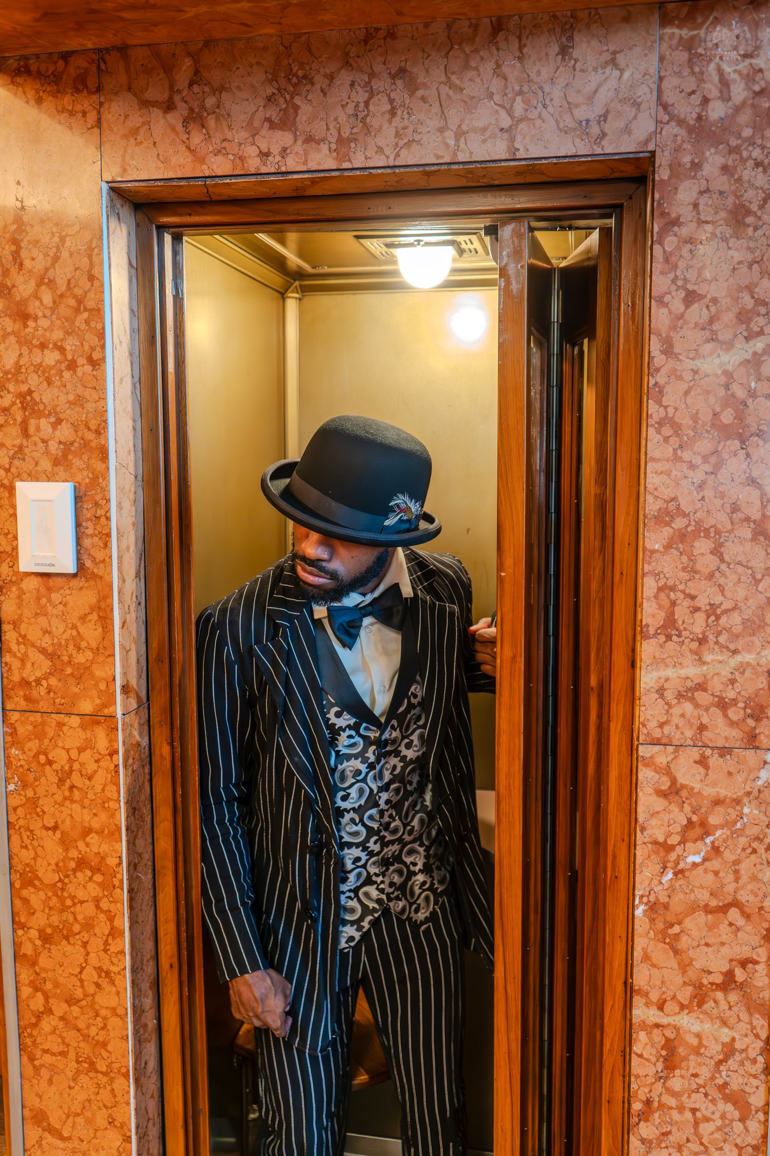 Stylish man in pinstripe suit, bowler hat, and bow tie posing inside an elegant elevator with marble walls.
