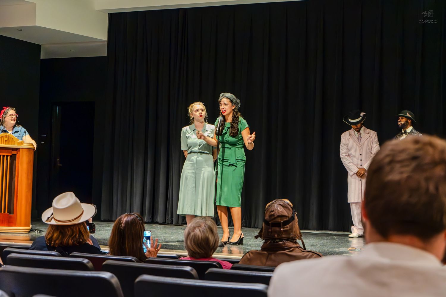 Two women in vintage green and blue dresses perform on stage during a theatrical production.