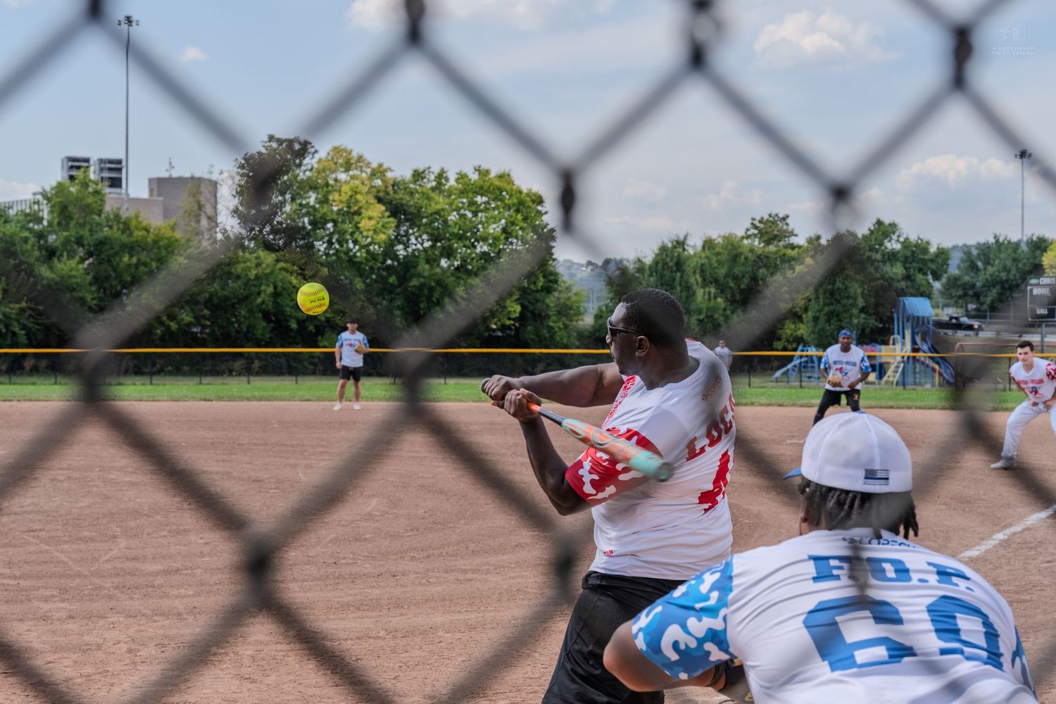 Softball batter swinging at a yellow ball viewed through a chain-link fence during an outdoor game.
