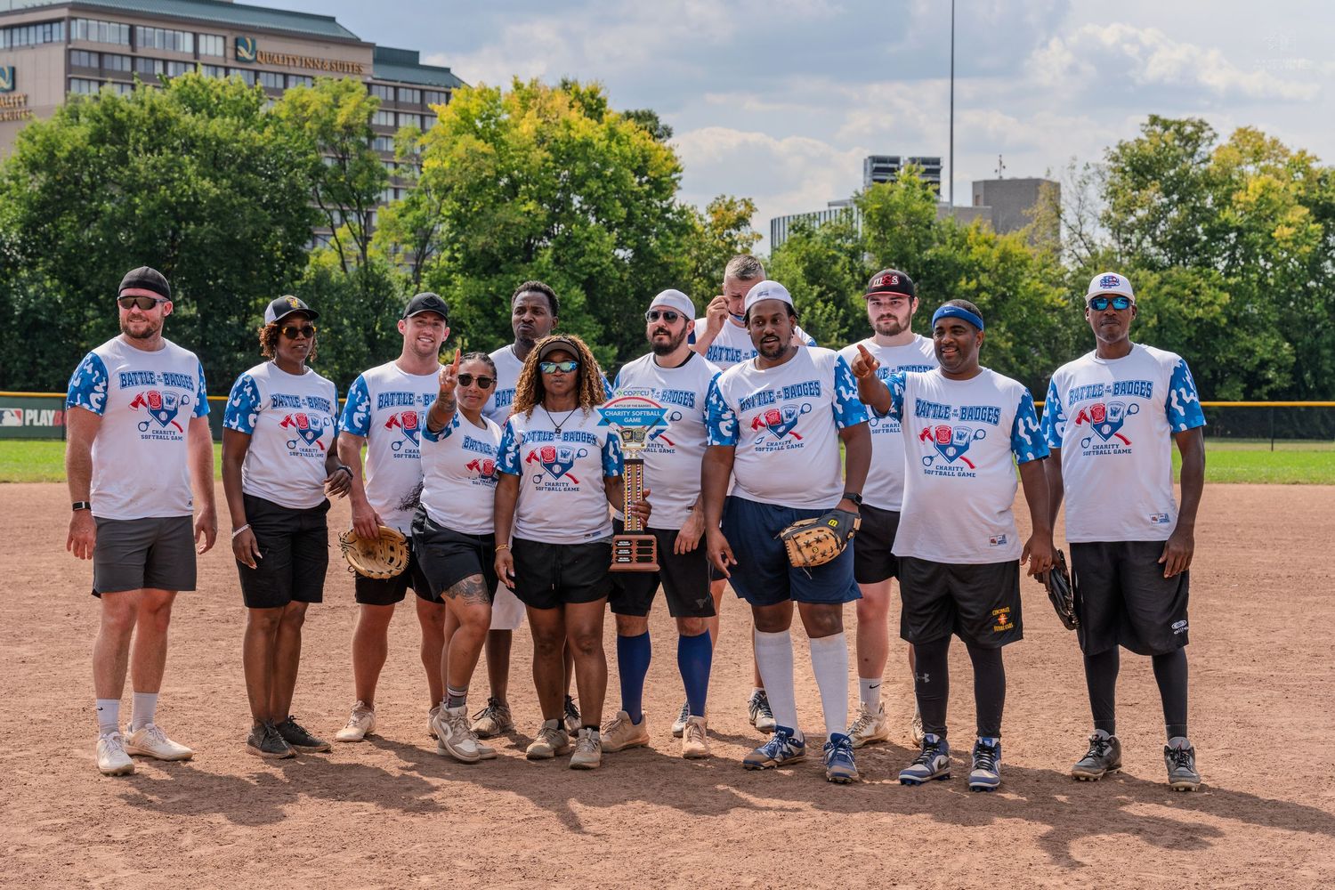 A softball team poses together on a field in matching white jerseys, celebrating with a trophy on a sunny day.