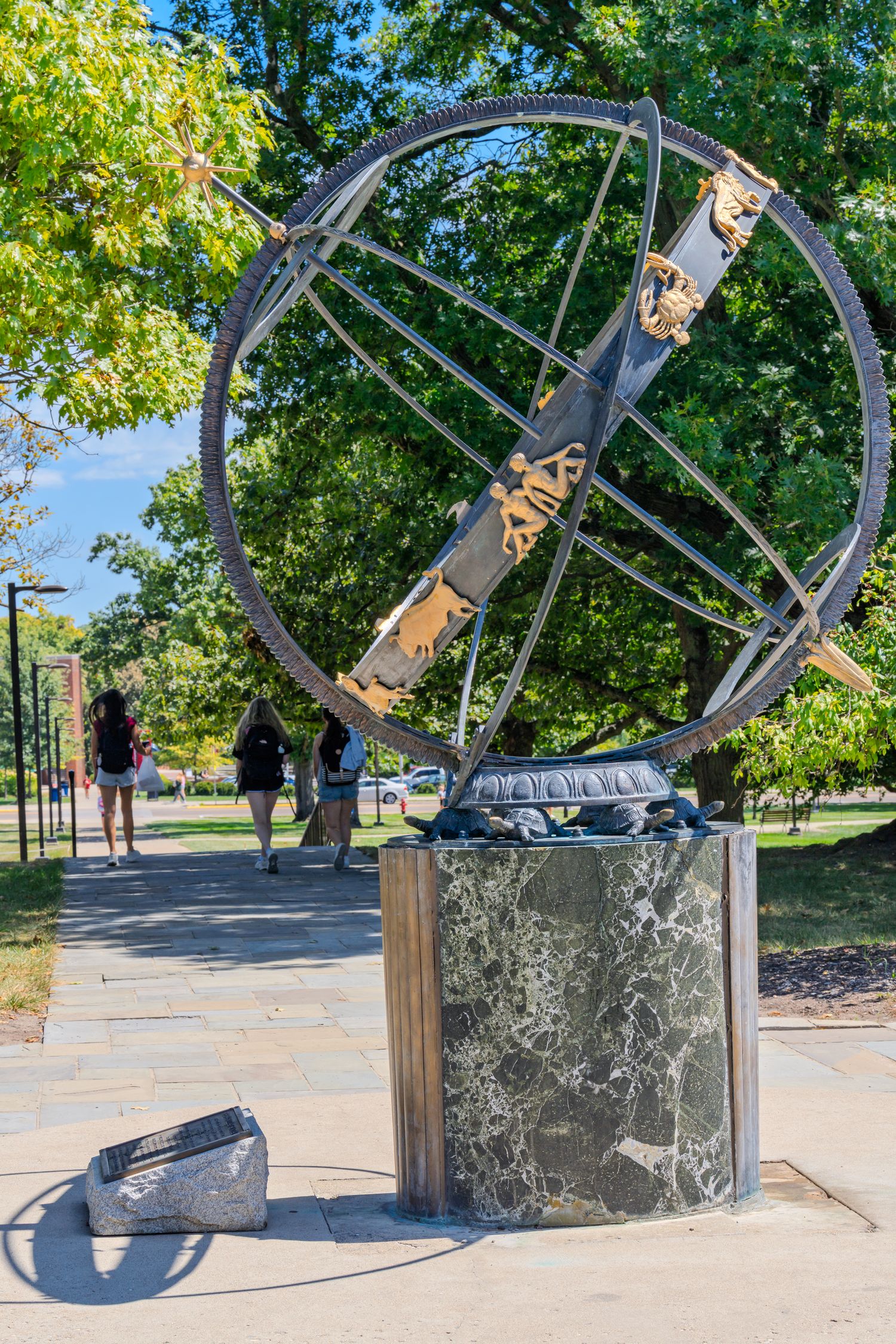 Armillary sphere sundial on marble pedestal in a park with tree-lined path and visitors in background.