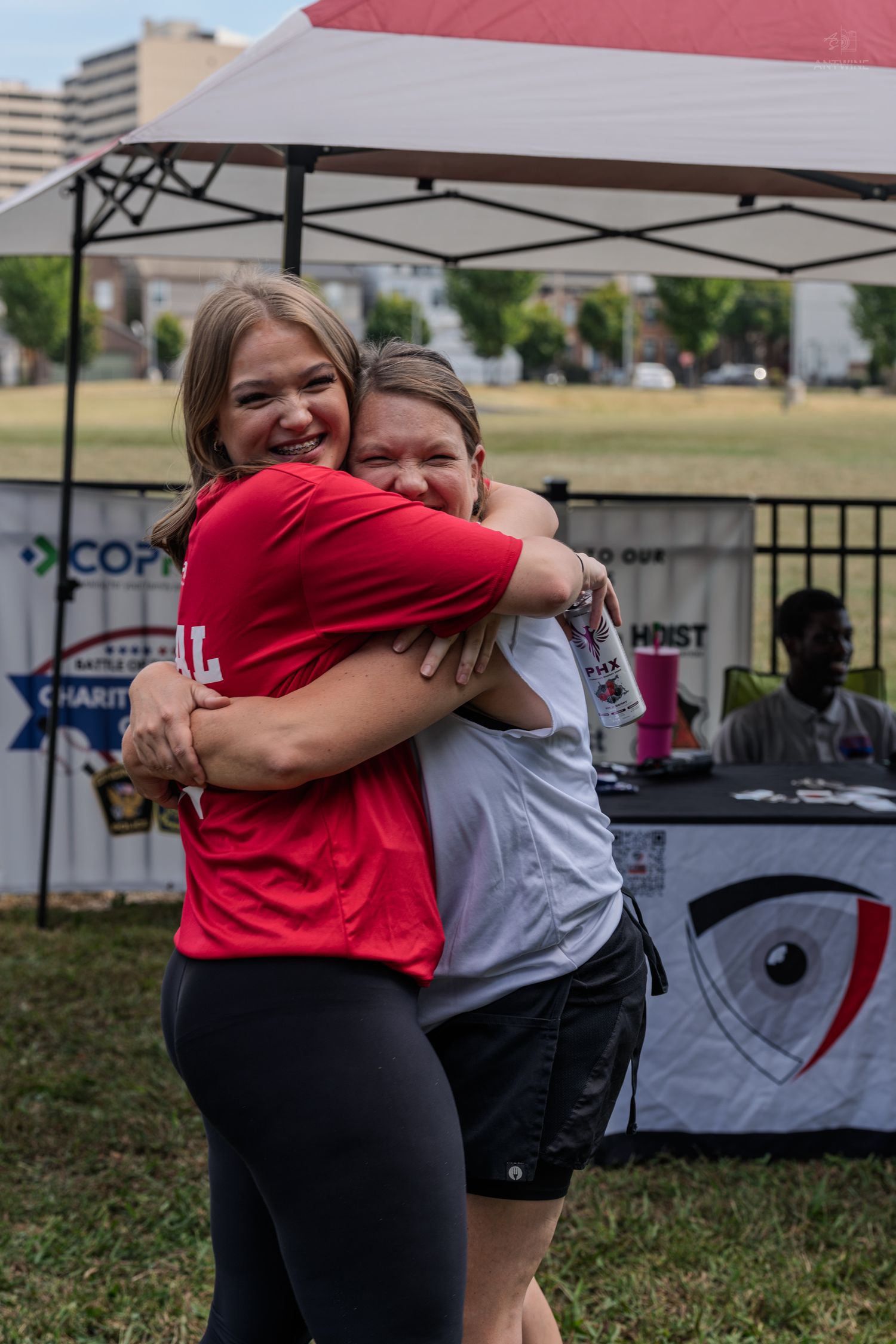 Two women sharing a warm hug at an outdoor charity event under a white canopy tent on a grassy field.