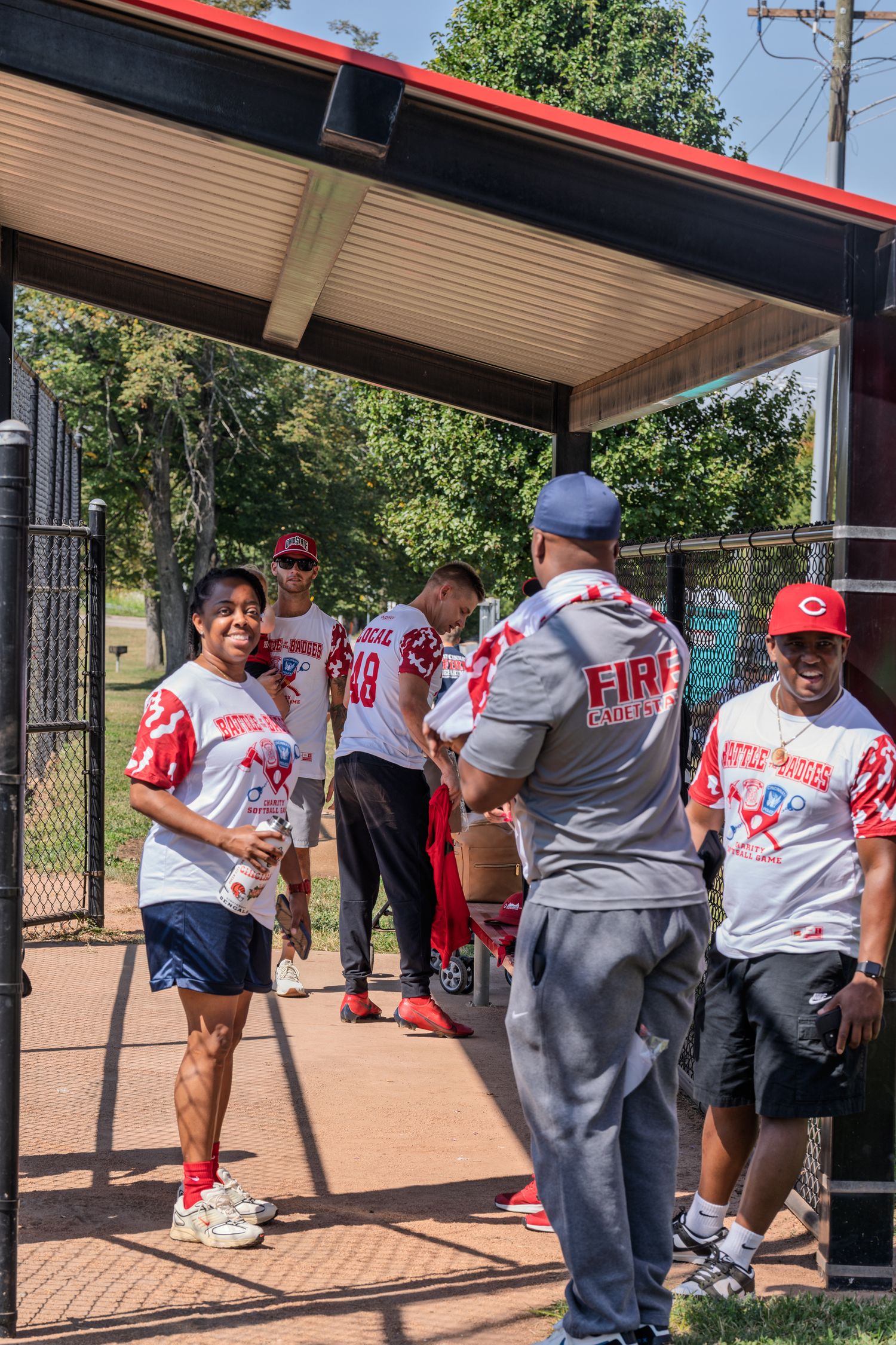 Fire department softball team members in red and white uniforms gathered in dugout area on sunny day.