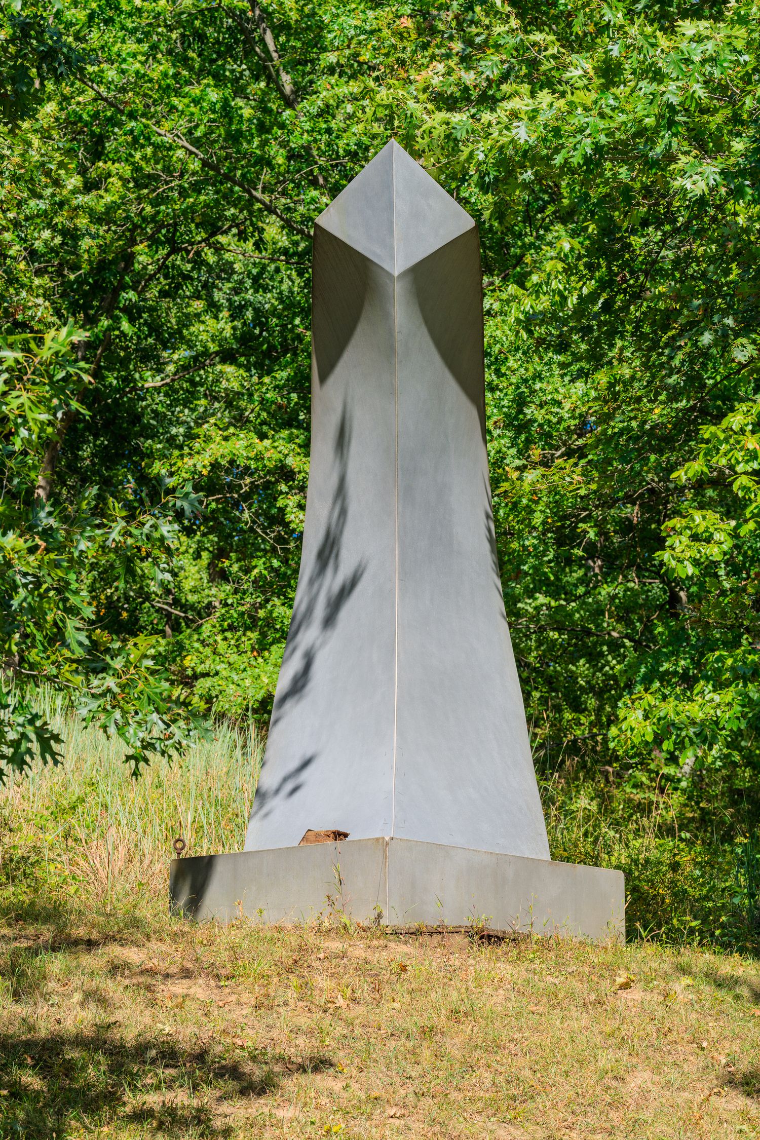 Modern abstract metal sculpture on concrete base surrounded by lush green trees in an outdoor park setting.