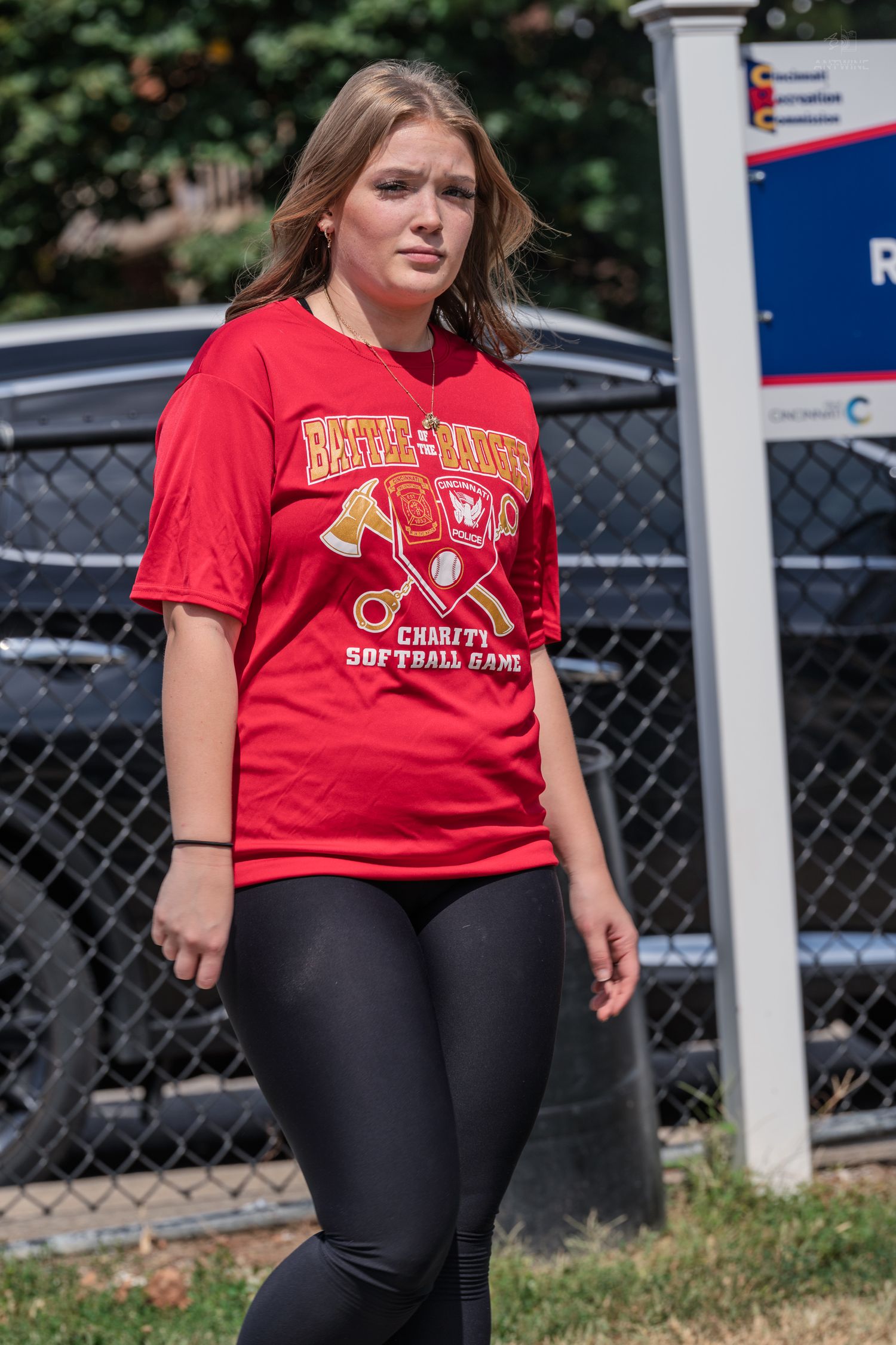 Young woman in red charity softball game t-shirt and black leggings walking outdoors.