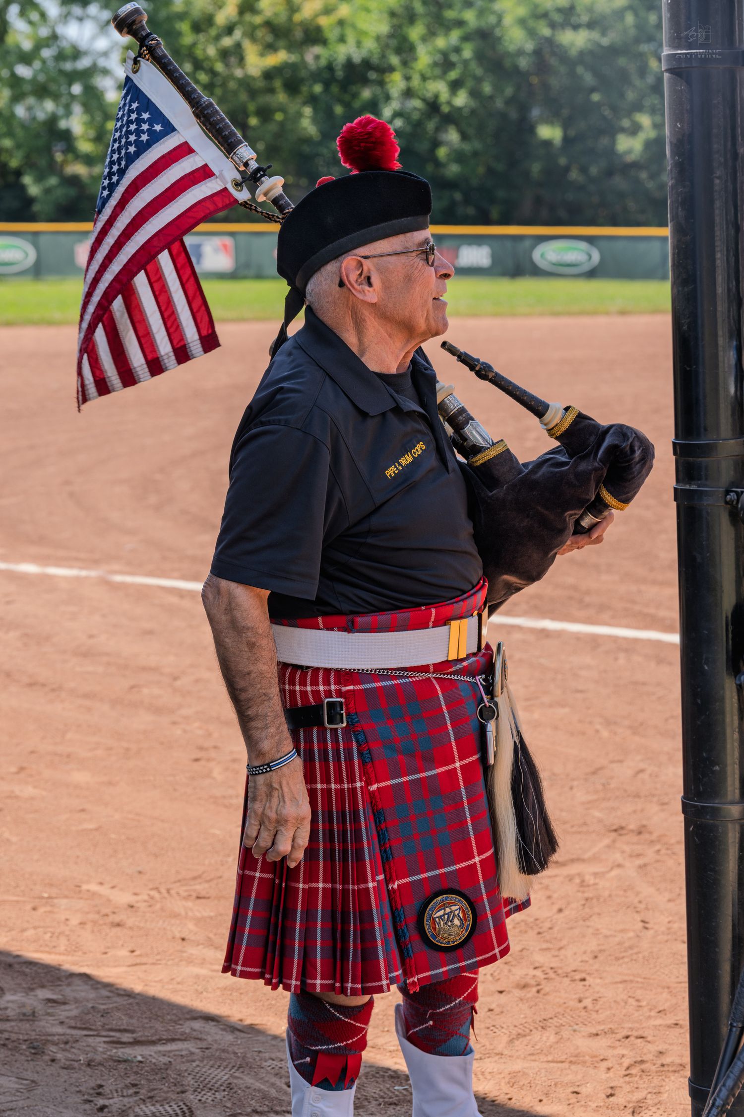 A bagpiper in a red tartan kilt plays beside an American flag pole on a baseball field.