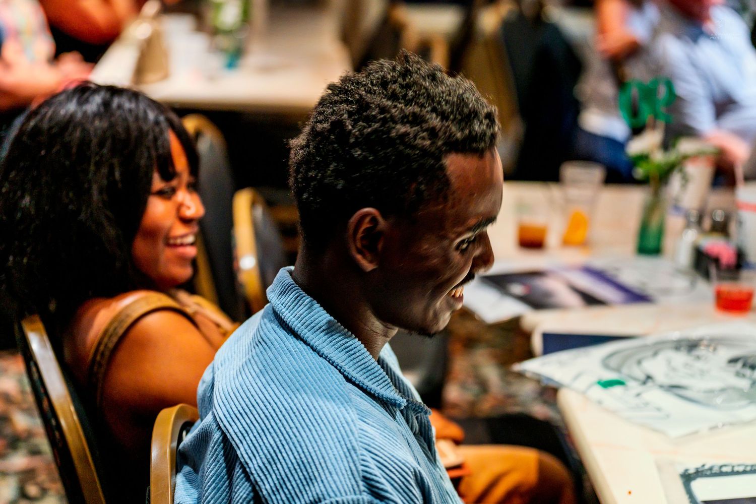 A young Black man smiles while seated at a table during an indoor event, with a laughing woman visible in the background.