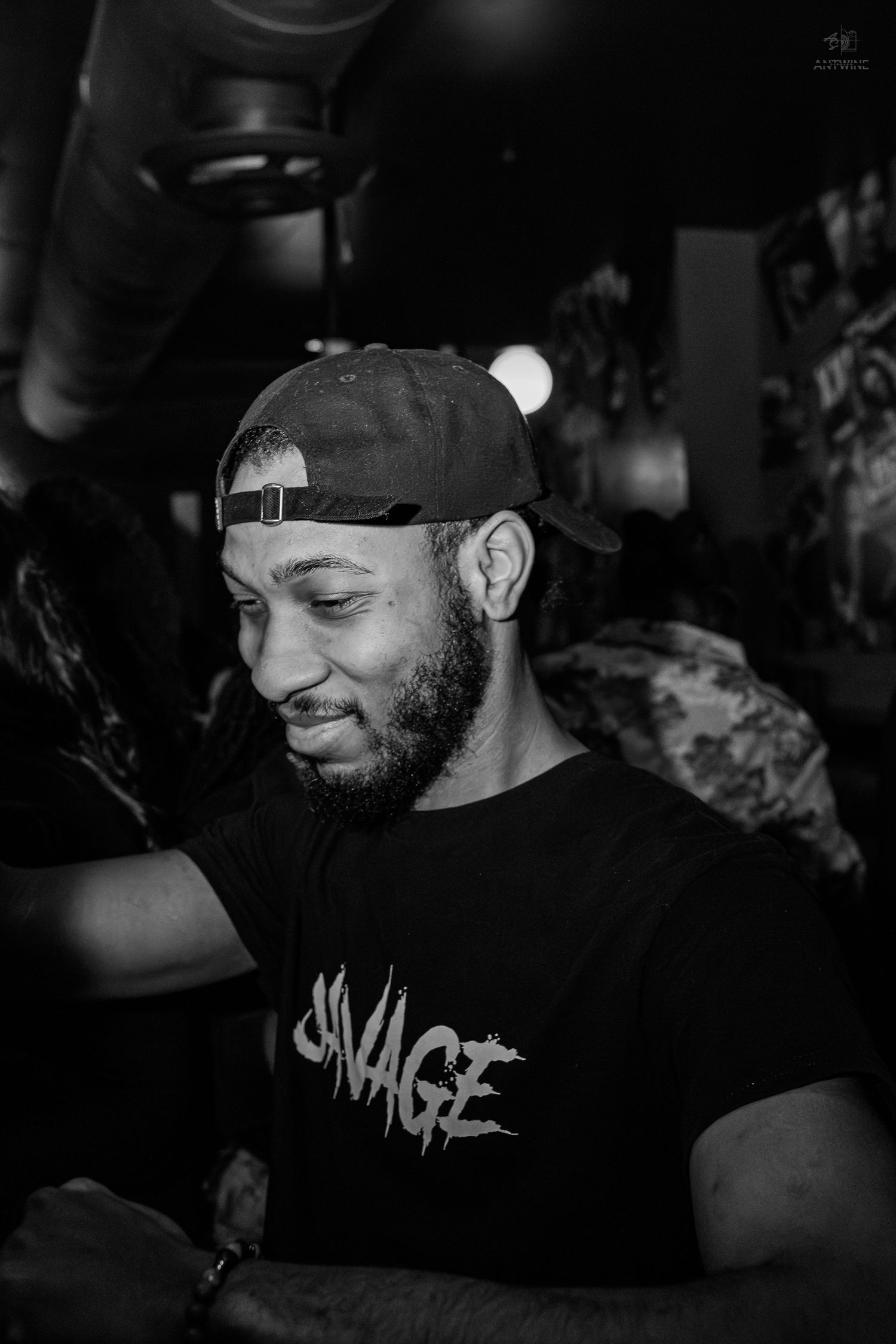 Man wearing a backwards cap and Savage t-shirt smiling in a dark venue, black and white photo.