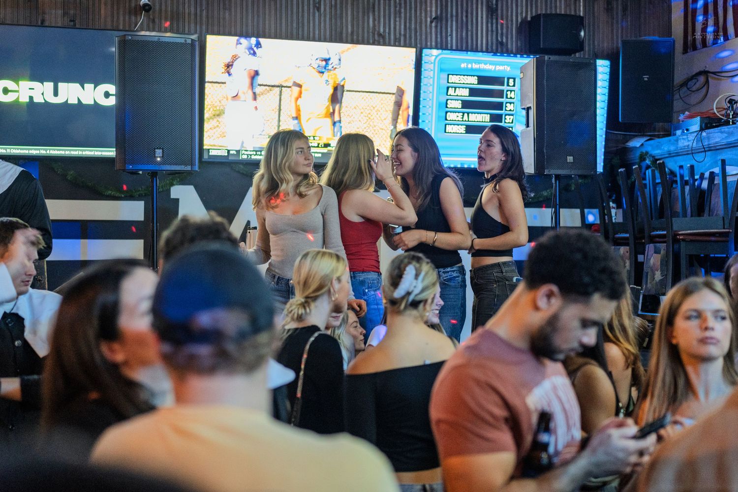 Group of women dancing and socializing at a lively sports bar with large TV screens in the background.