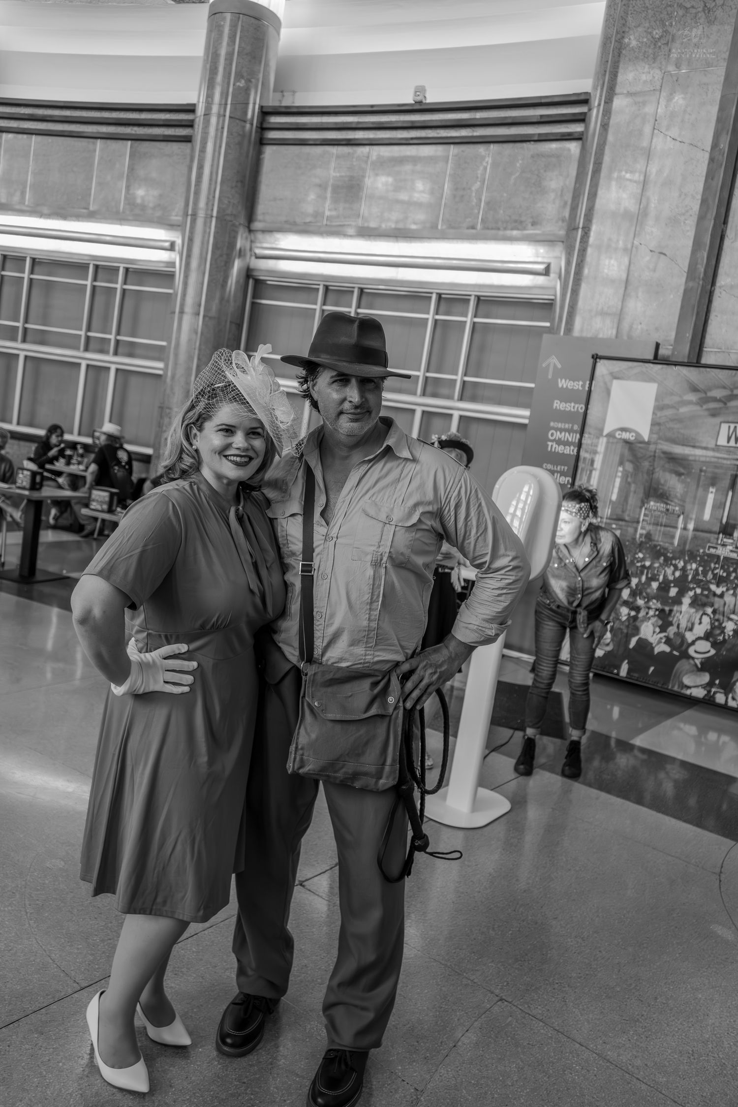 Black and white photo of a couple in vintage 1940s costumes posing in a grand hall at a themed event.