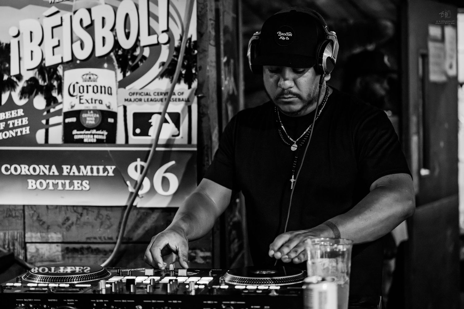 Black and white photo of a DJ wearing headphones mixing at turntables near Corona beer signs.