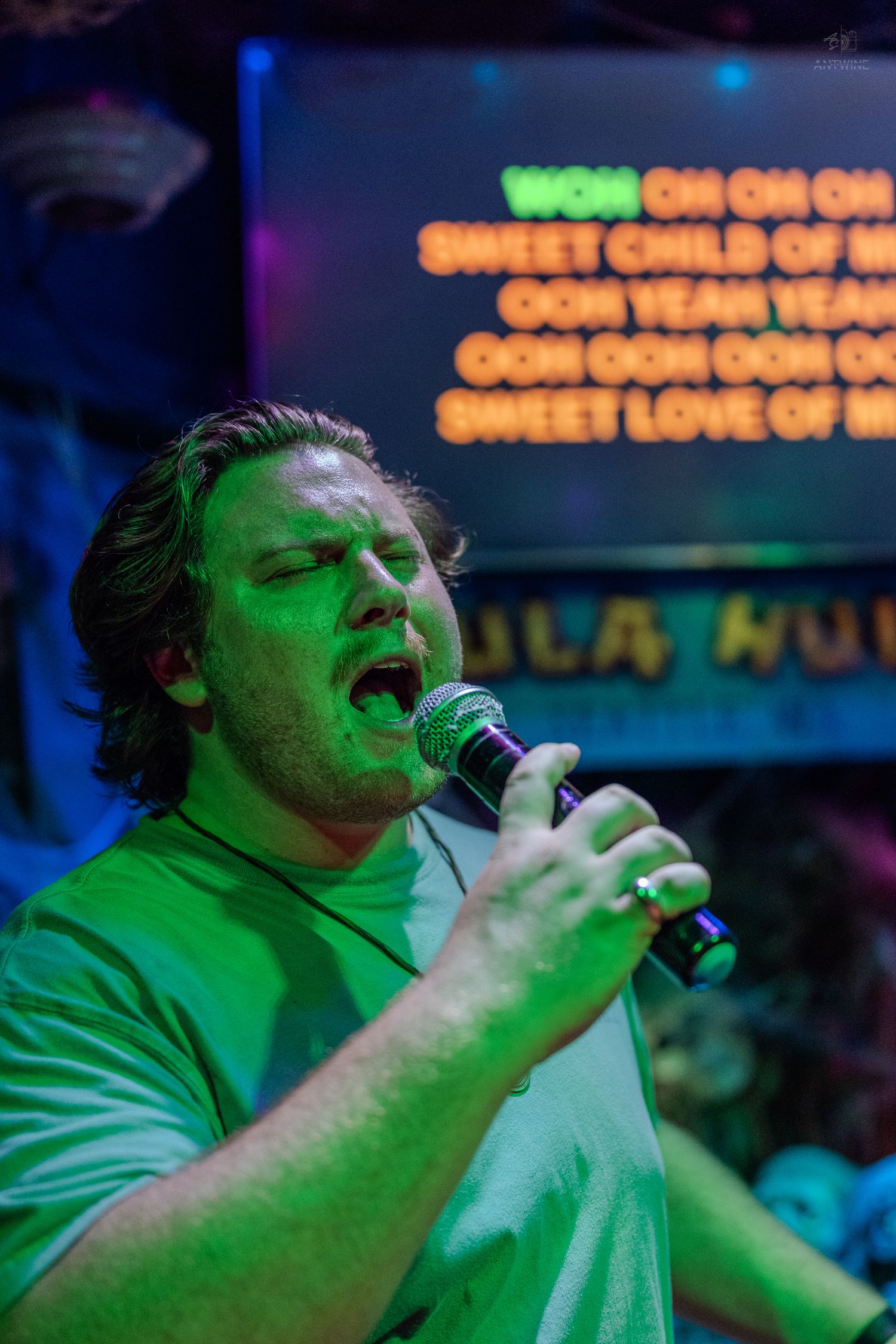 Man singing karaoke passionately into microphone under green stage lights with Sweet Child O Mine lyrics on screen.