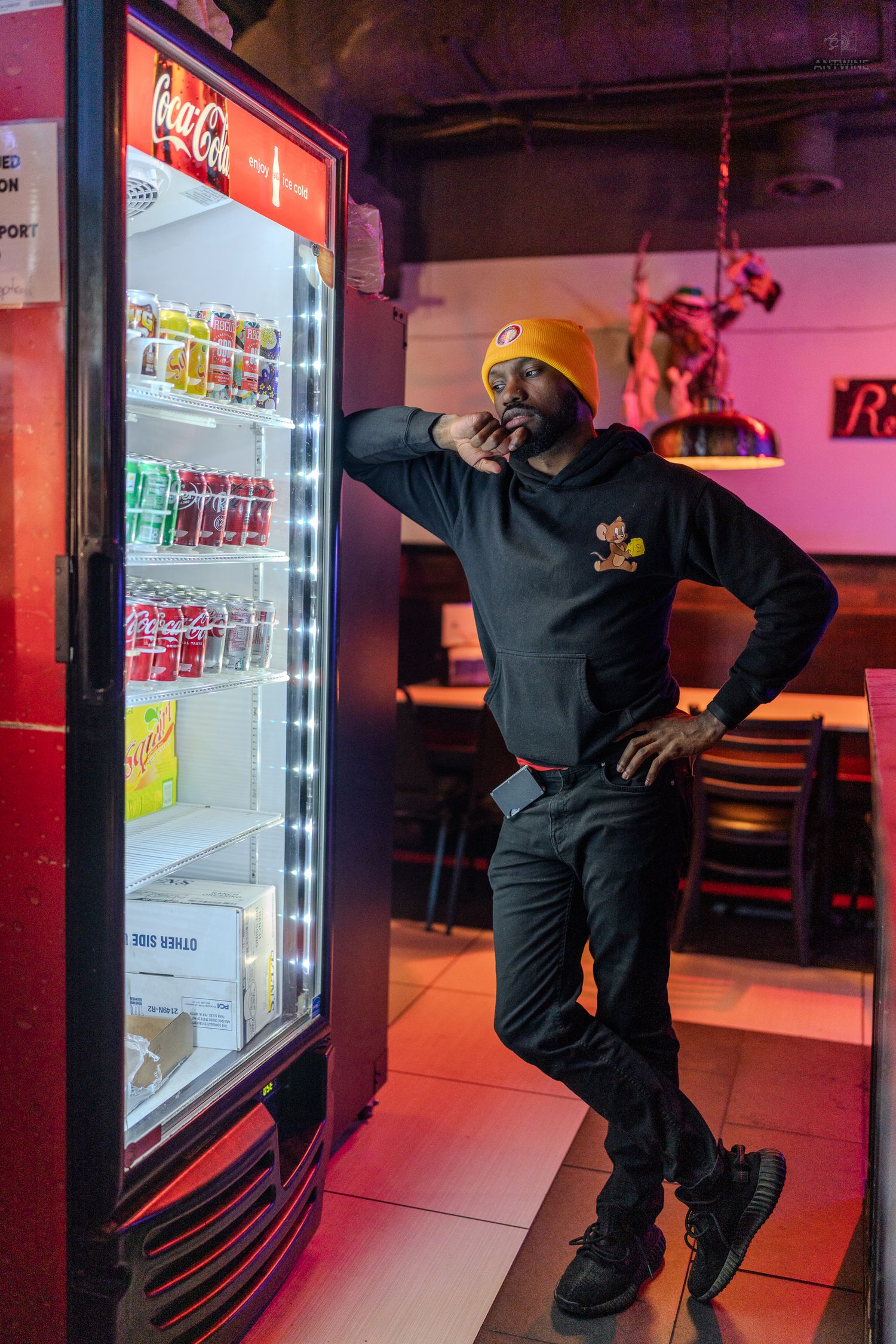 Man in black hoodie and orange beanie leaning against illuminated Coca-Cola refrigerator in a restaurant.