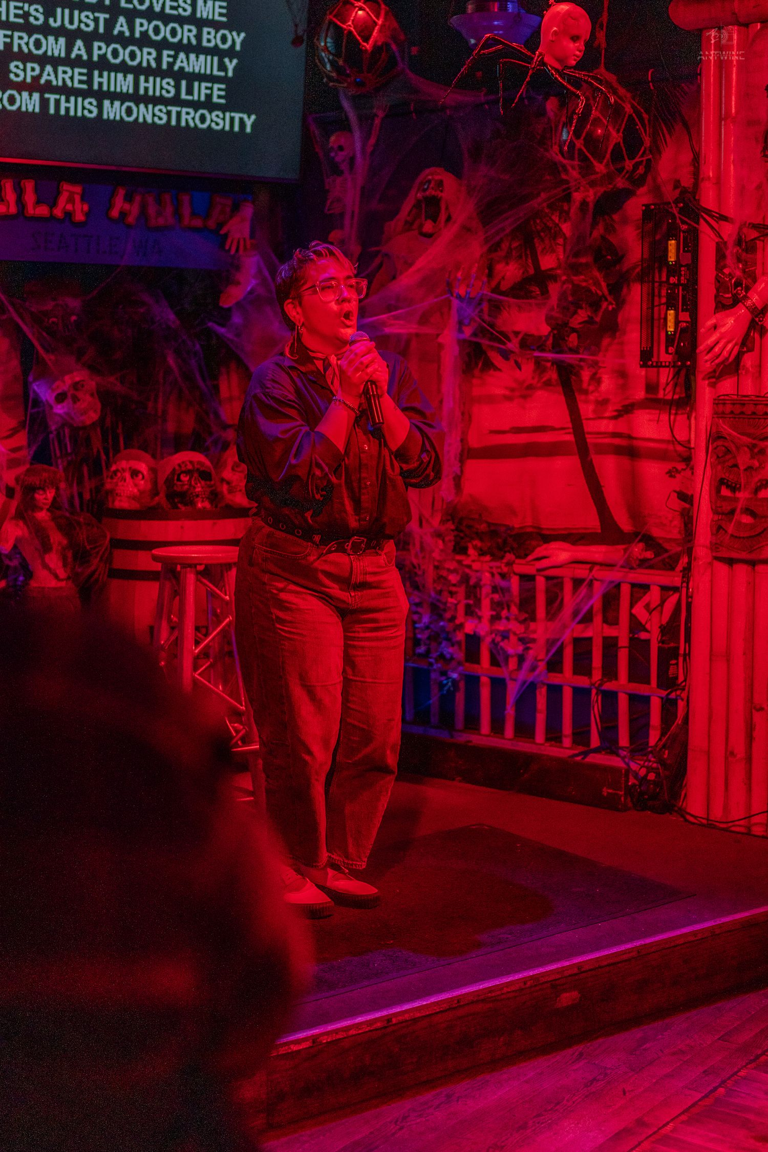 Person singing karaoke on Halloween-decorated stage bathed in red light at Hula Hula bar.