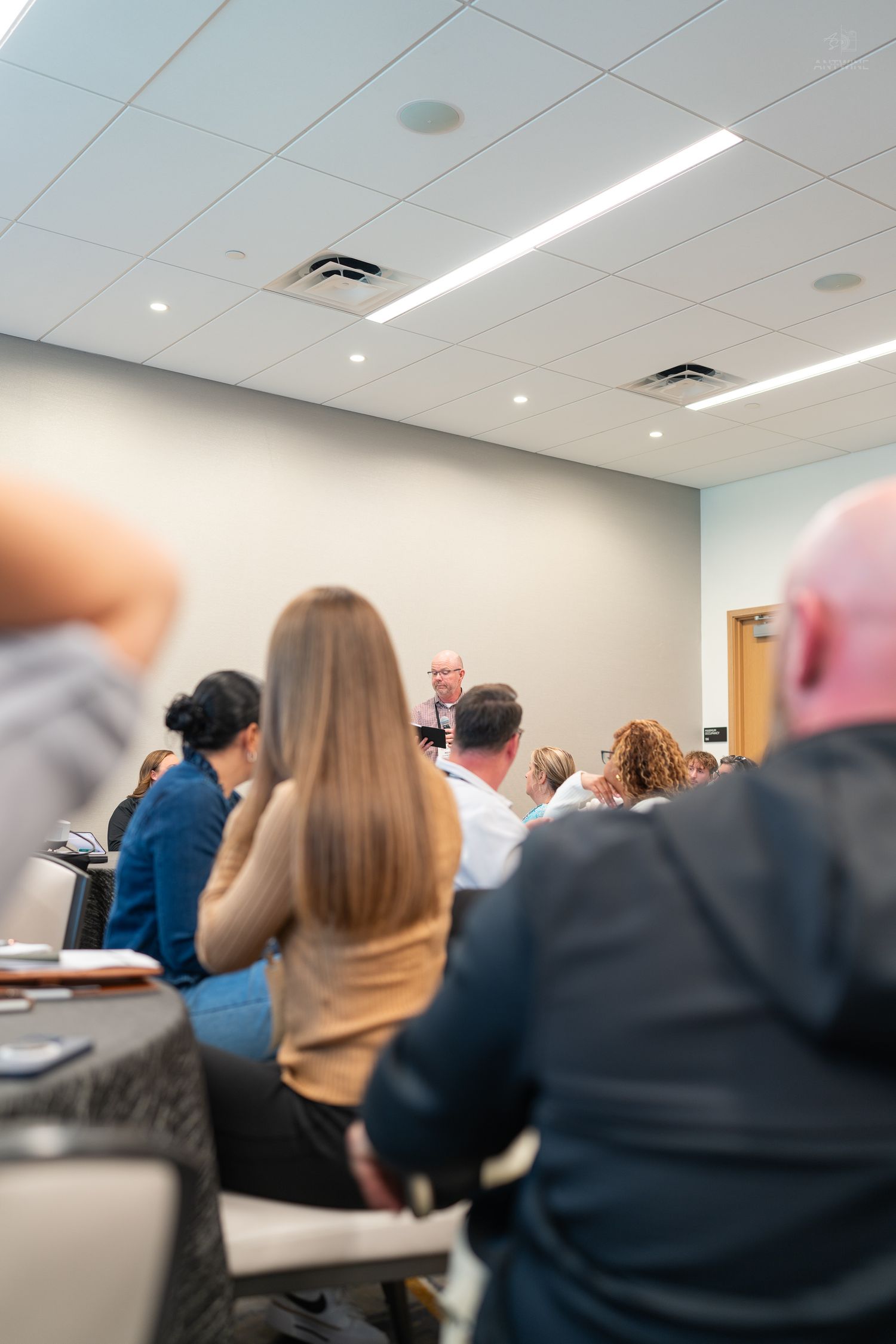 Audience seated at a professional conference listening to a speaker at the front of a bright room.