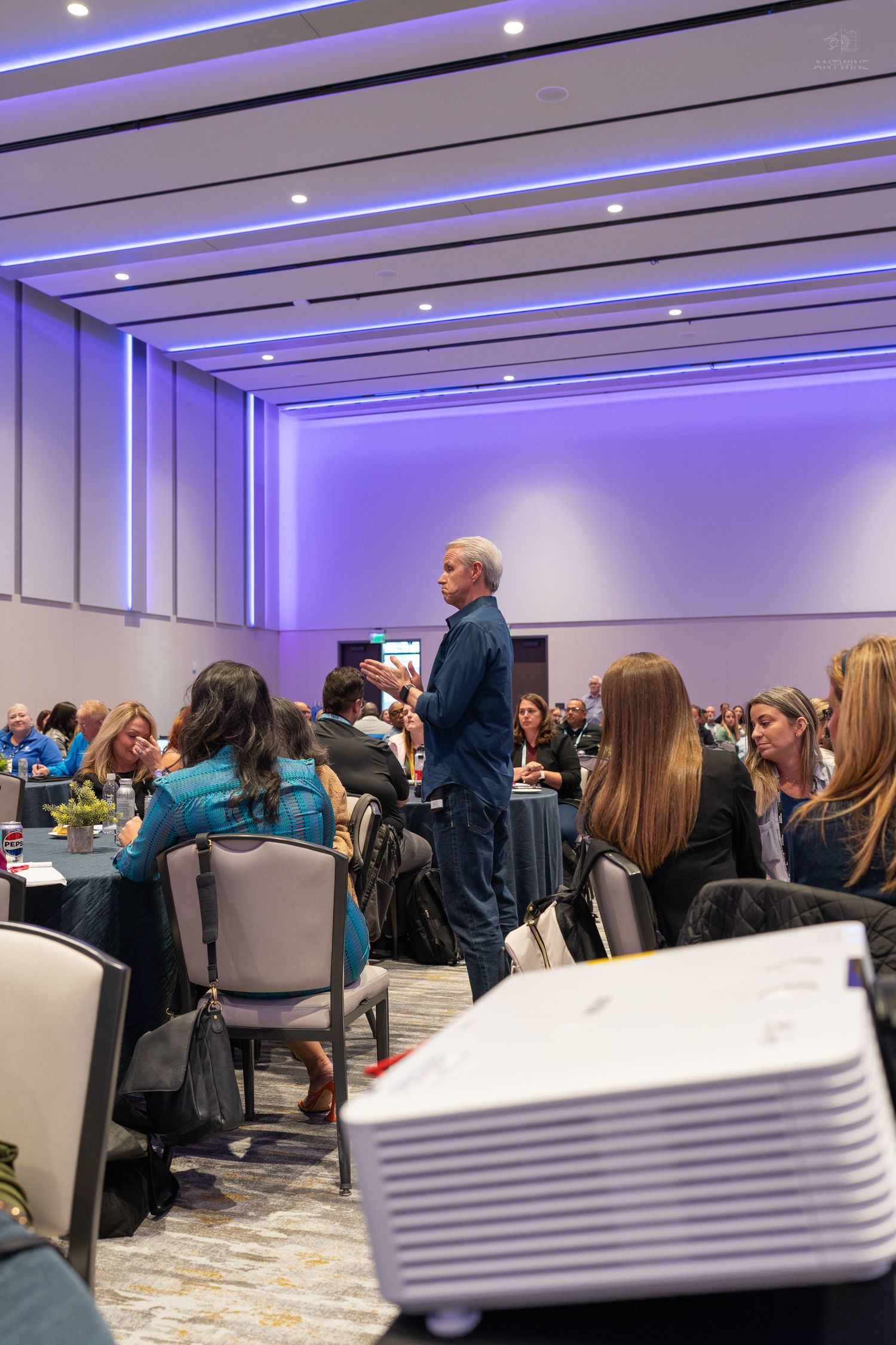 A presenter addresses a large audience seated at round tables in an elegant ballroom with purple lighting.