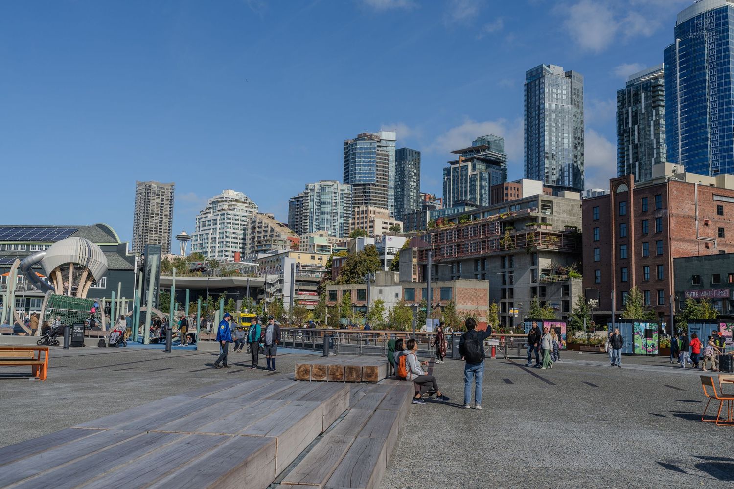 Seattle waterfront plaza with modern skyscrapers, historic brick buildings, and people enjoying the outdoor public space.