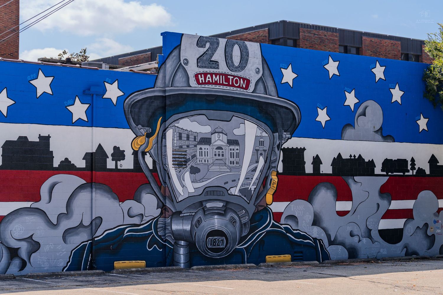 Firefighter mural on brick wall with American flag design, helmet reading Hamilton 20, and gas mask in urban setting.