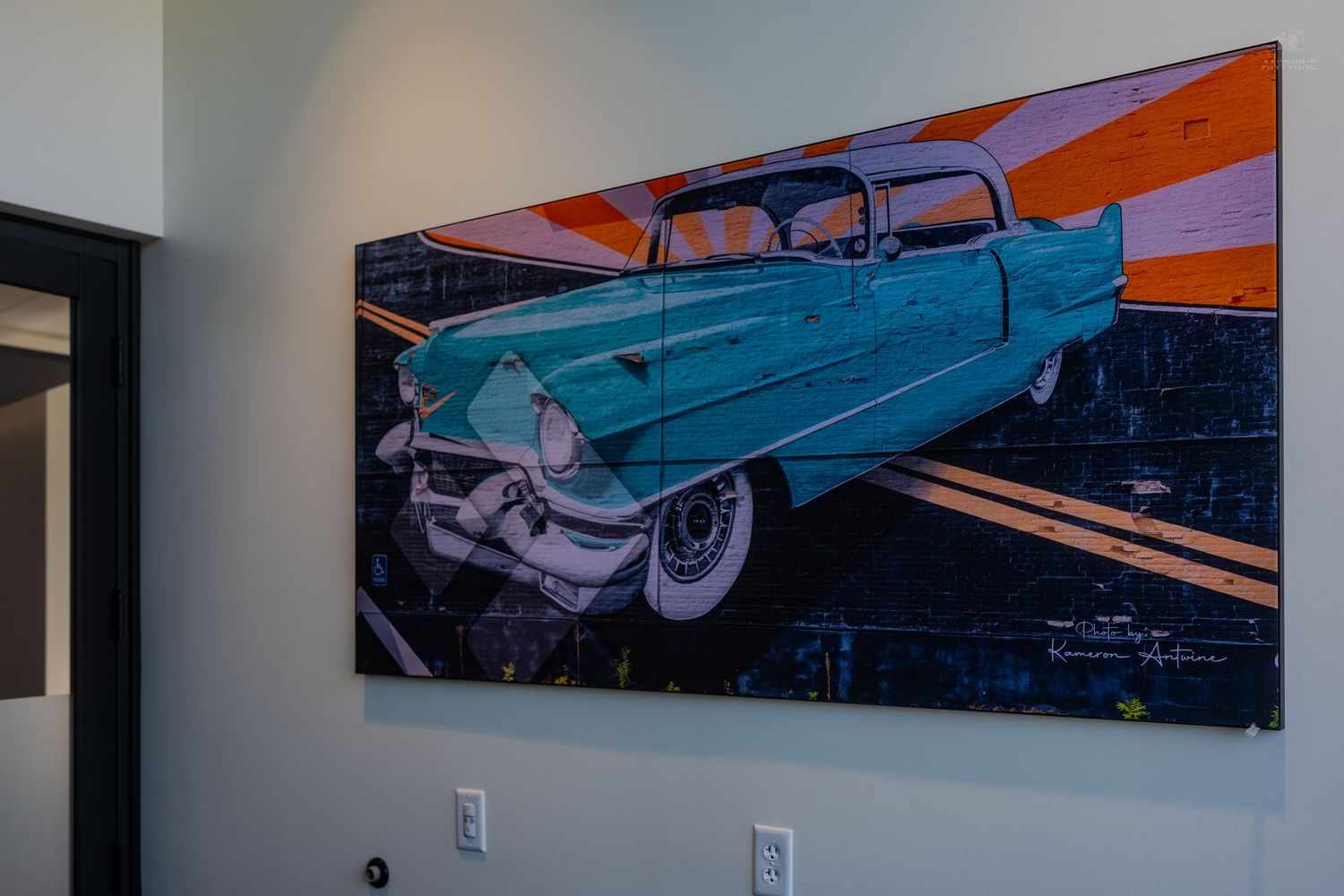 Pop art painting of a vintage teal Cadillac on an orange sunburst background, displayed on a white interior wall.