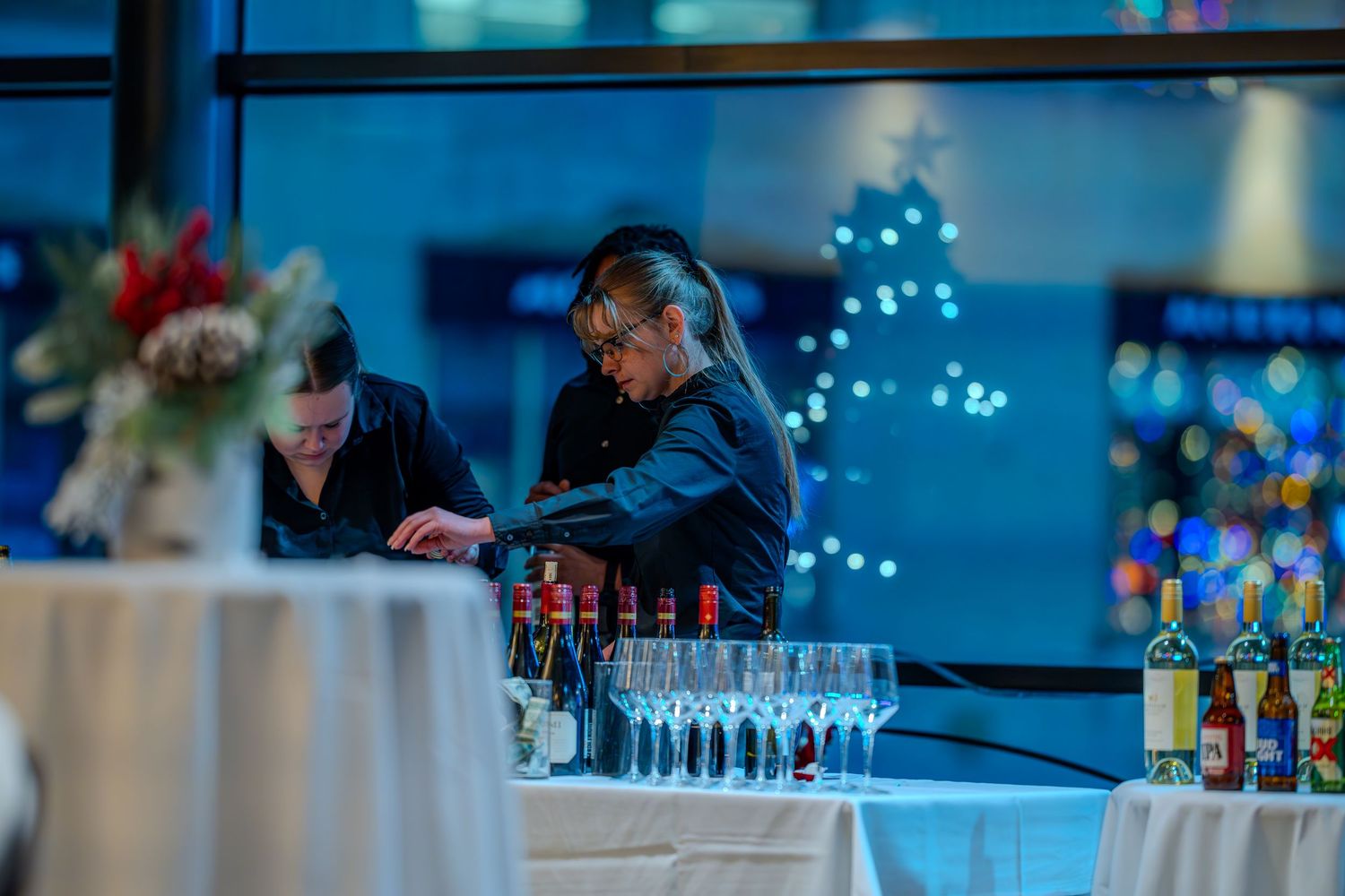Two bartenders prepare drinks at an elegant event bar with wine glasses and bottles, Christmas tree bokeh in background.