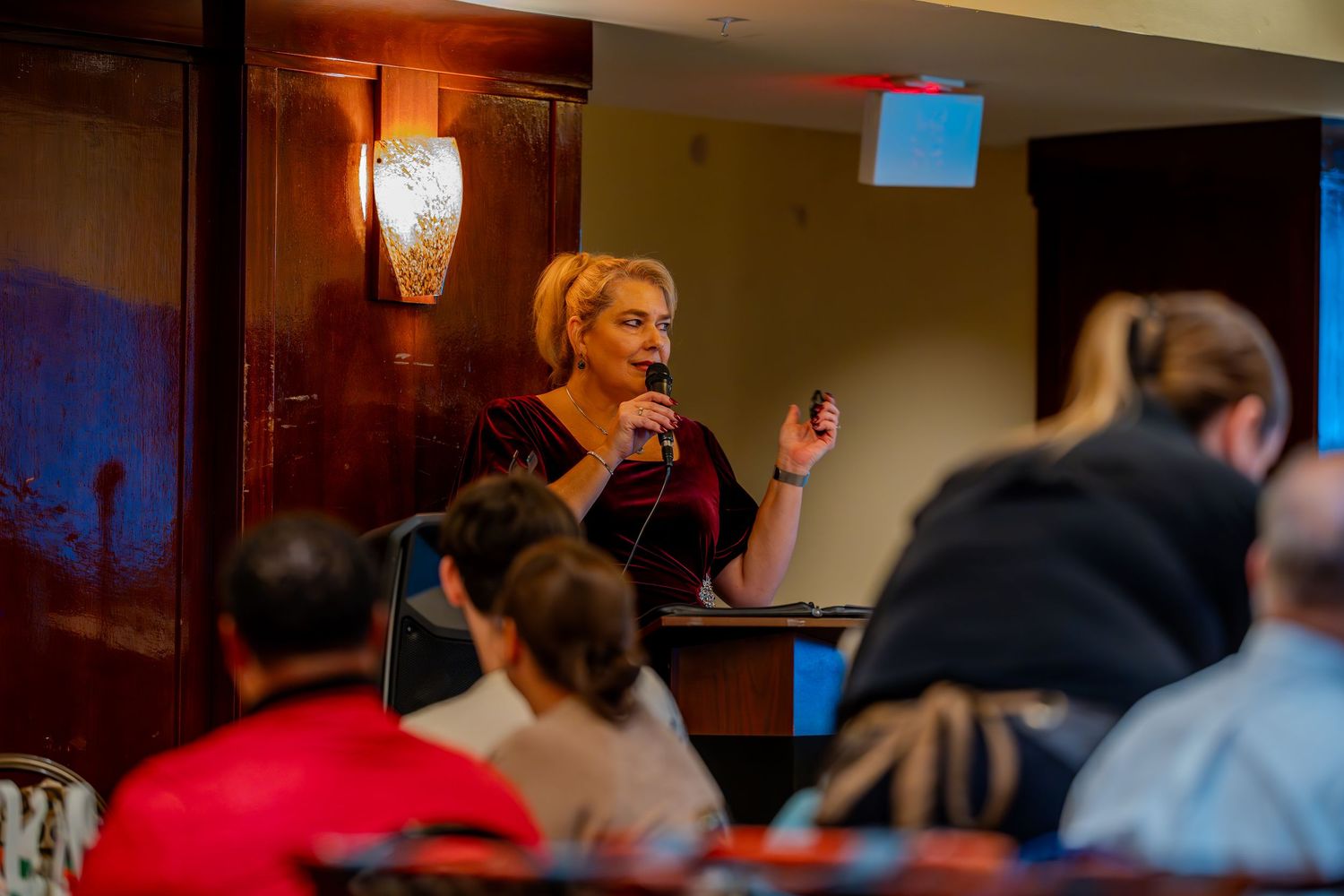 A blonde woman speaks at a podium with a microphone at an indoor event with an audience seated before her.