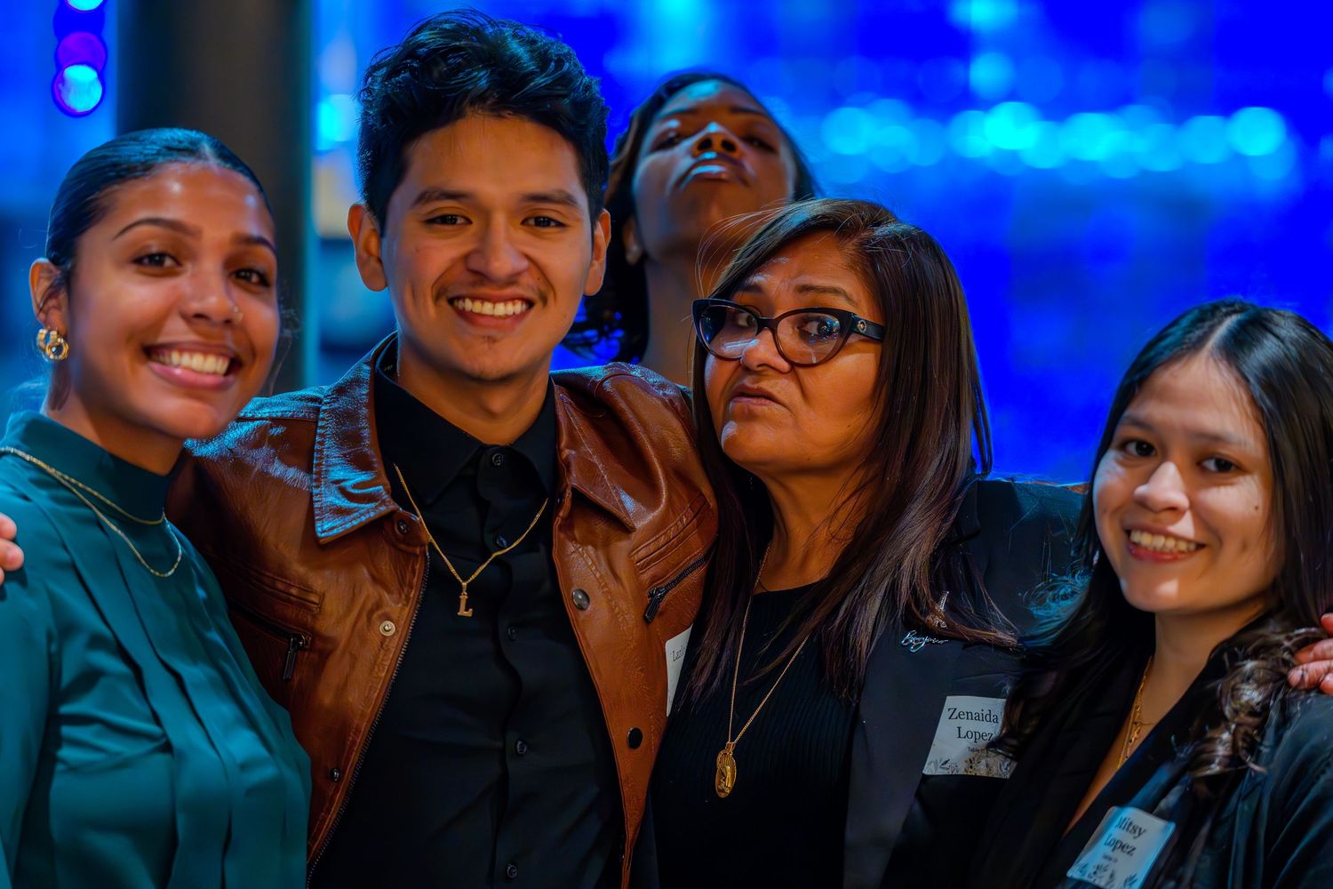 Five smiling young adults posing together at a formal event with blue ambient lighting in the background.