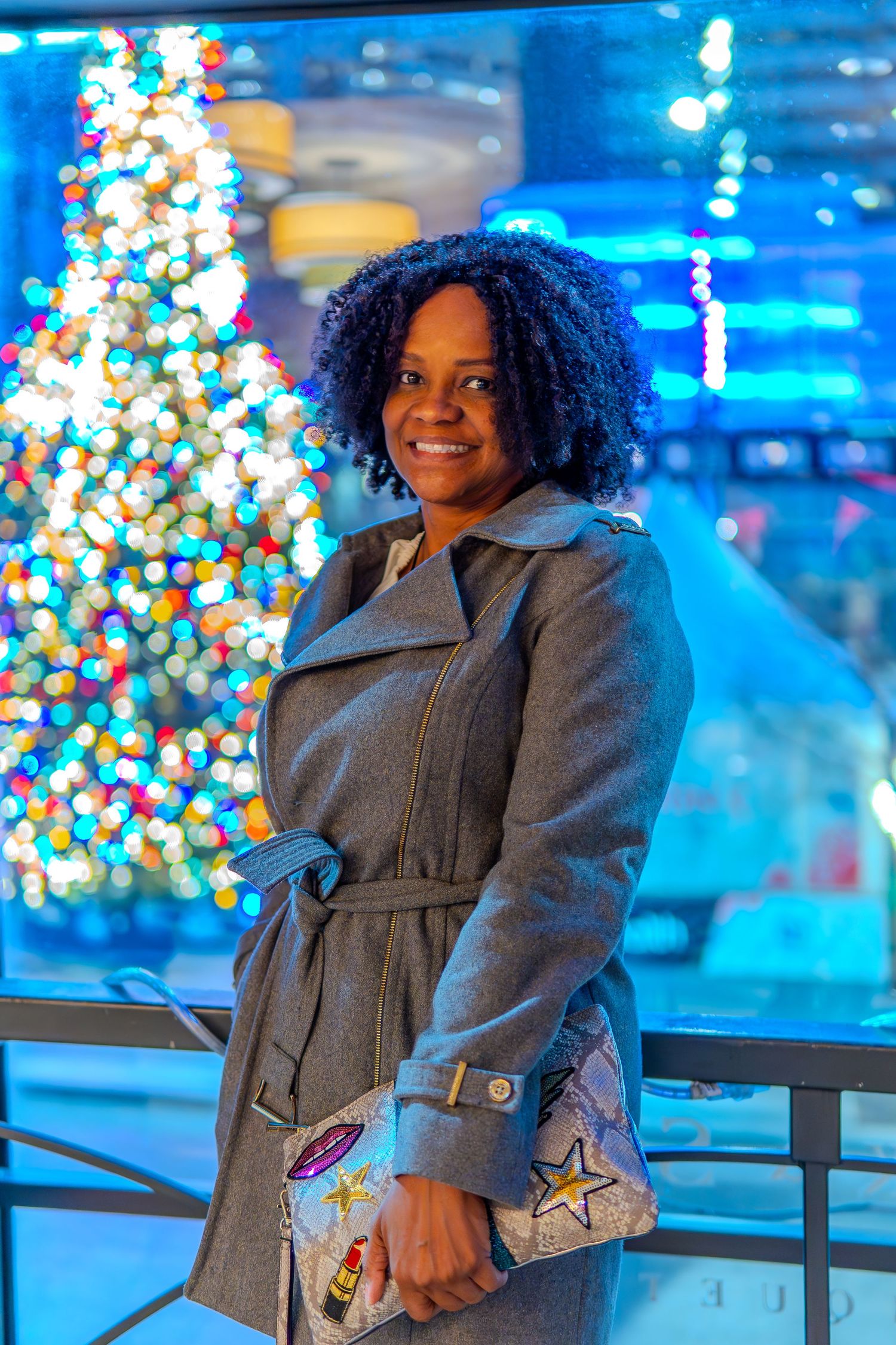 A smiling woman in a gray belted coat holds a designer clutch in front of a colorful Christmas tree with blue holiday lights.