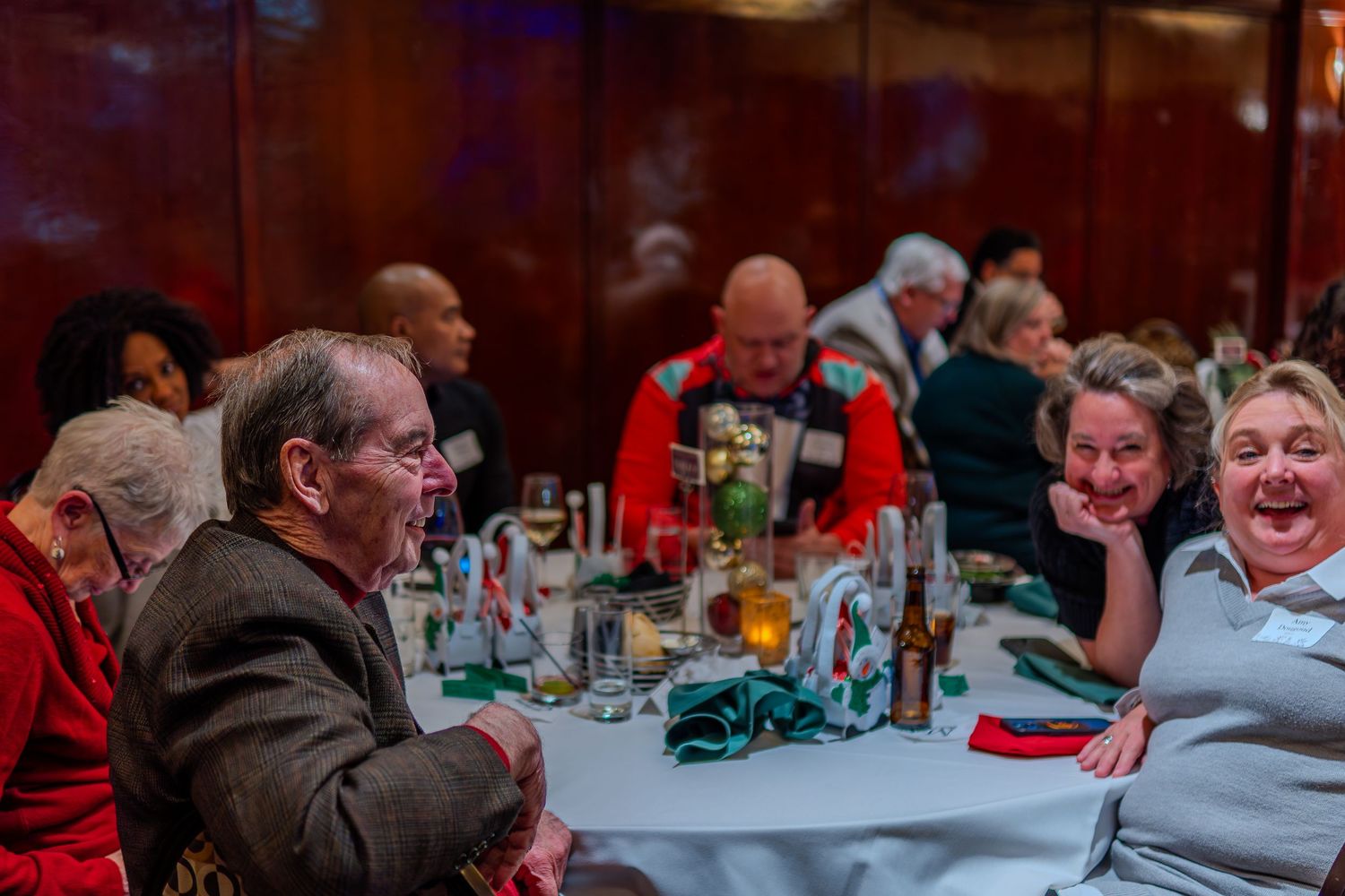 Group of people laughing and socializing at a festive dinner event, seated around a table with drinks and decorations.