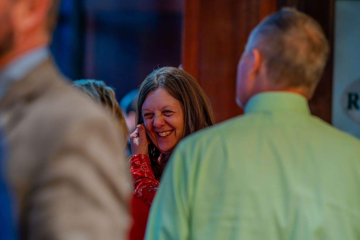 A woman in a red top laughs warmly at an indoor social gathering surrounded by guests.