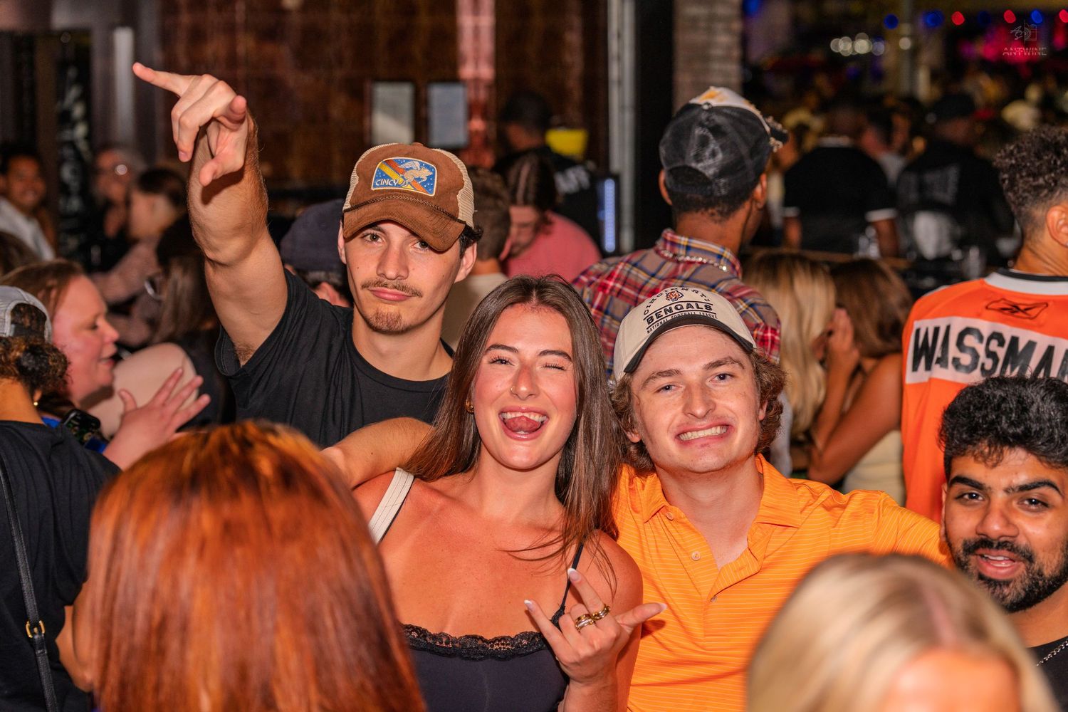 Three friends laugh and gesture together in a lively outdoor nighttime crowd event.