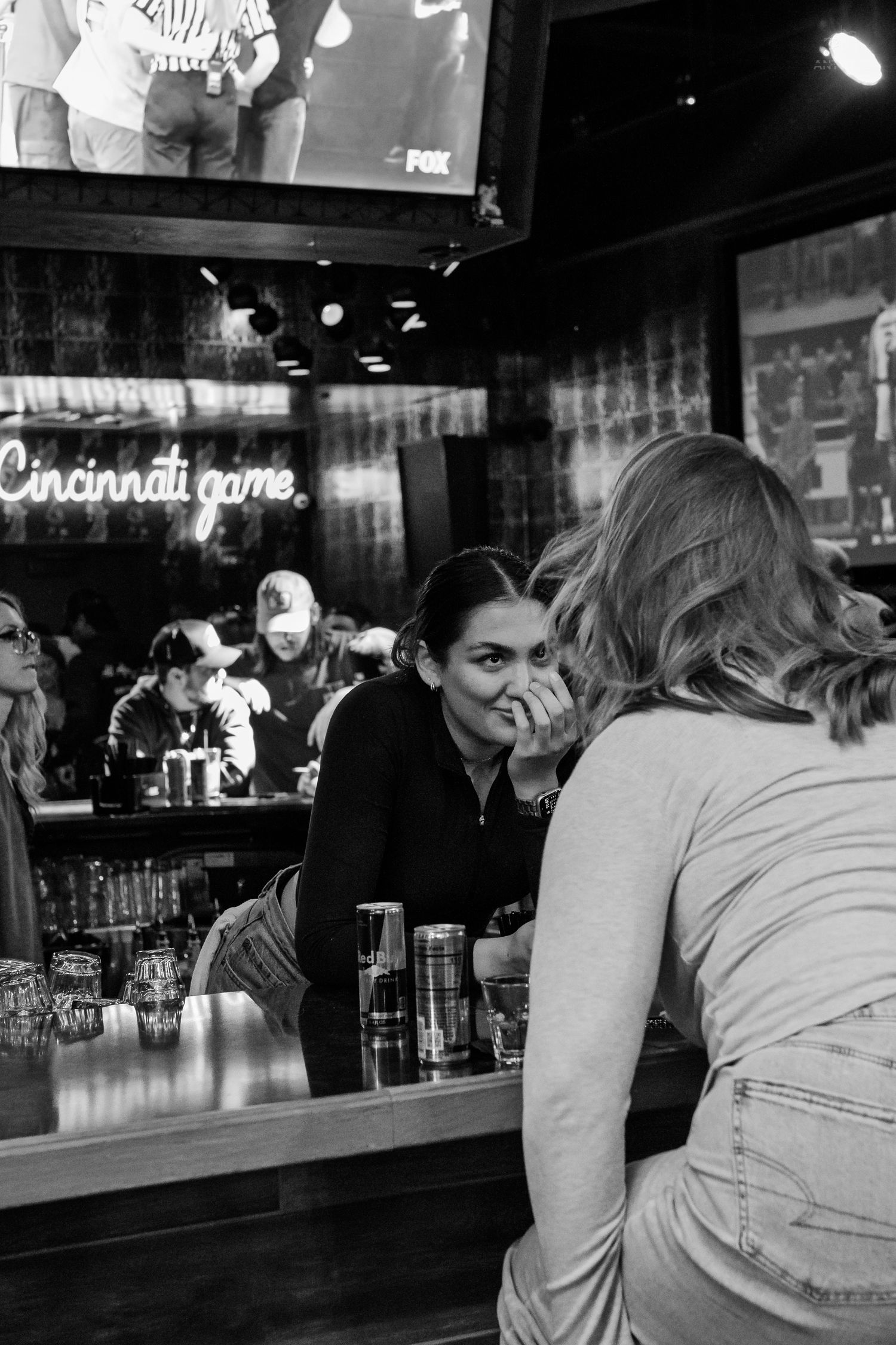 Black and white photo of two women talking at a bar counter with Cincinnati game neon sign behind.