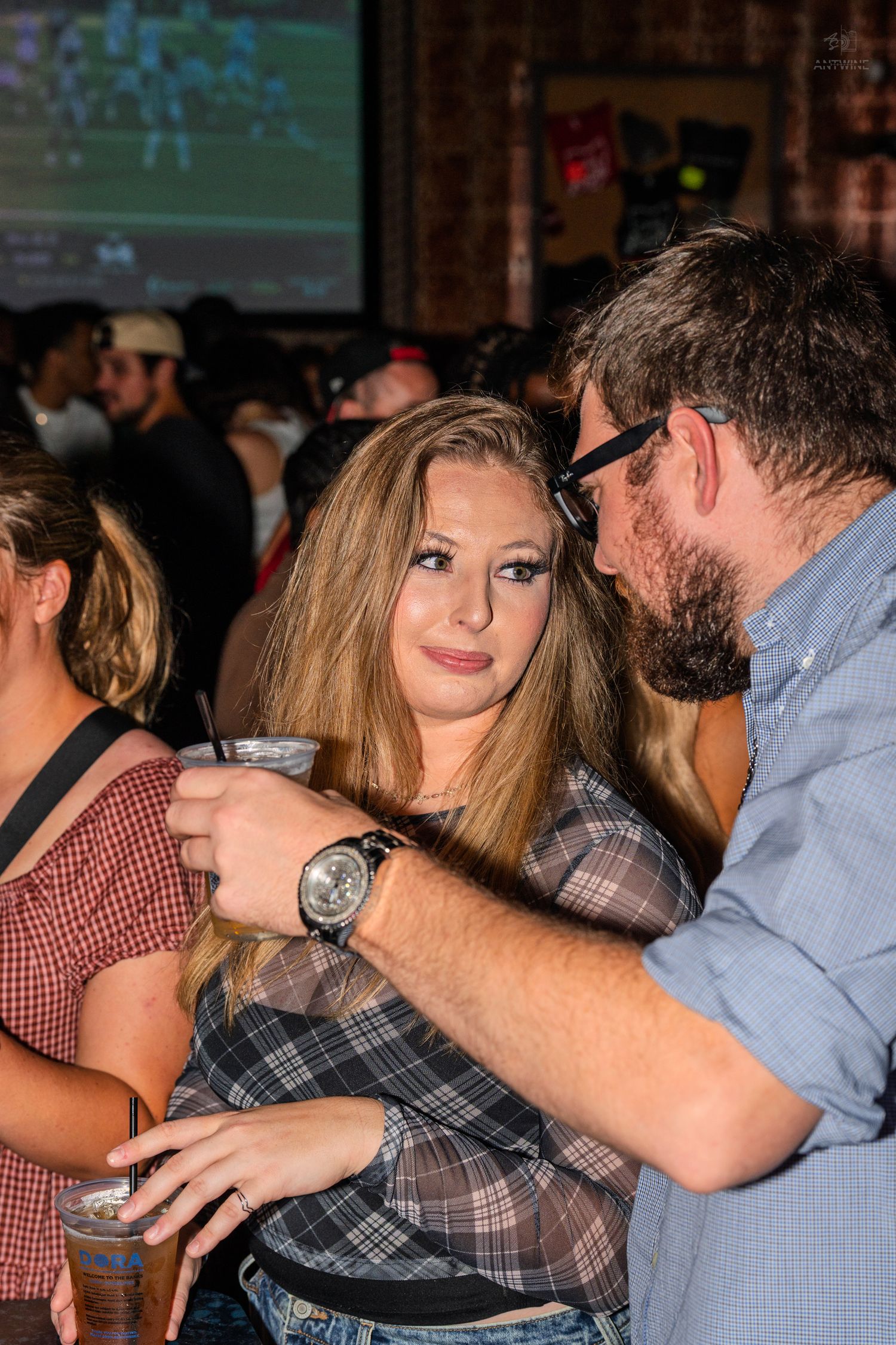 Young woman holding a drink while a bearded man leans in at a bar.