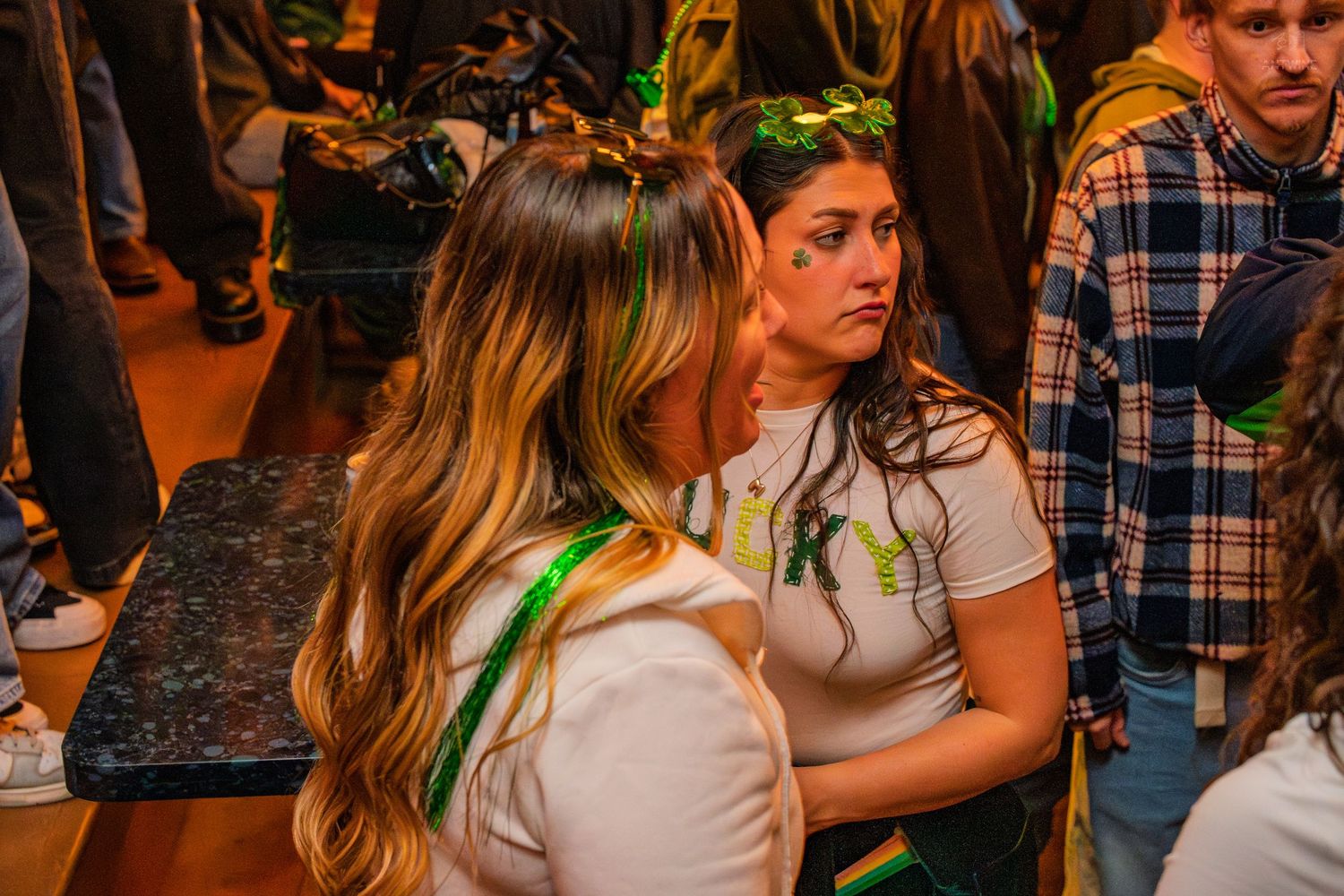 Two women in white shirts with green St. Patrick's Day accessories sitting together at a crowded bar.