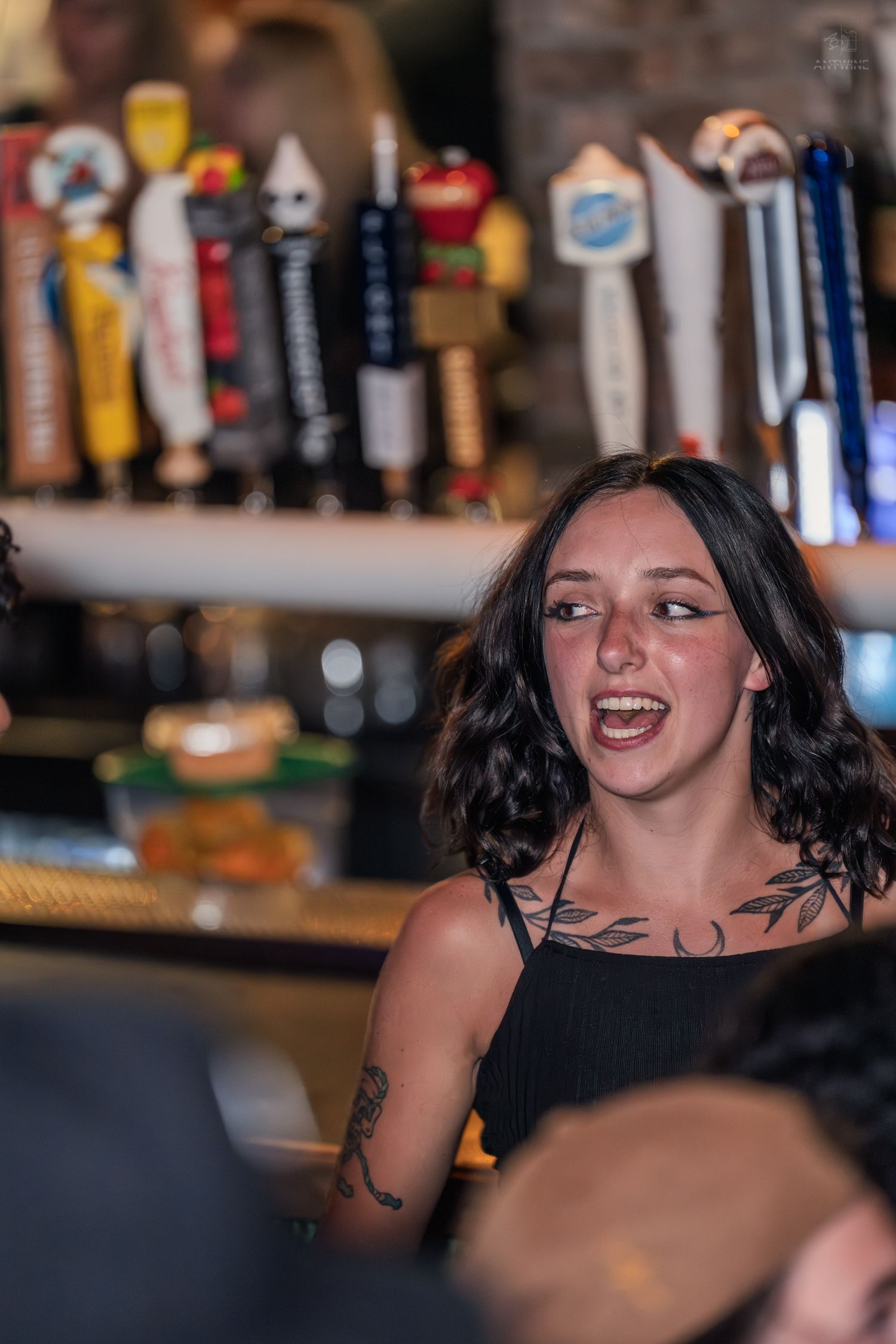 Smiling tattooed woman with dark wavy hair enjoying a lively conversation at a bar.