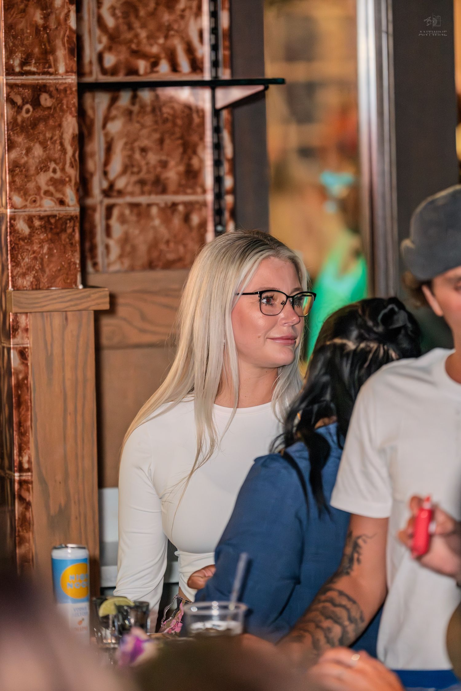 Blonde woman in white long-sleeve top and glasses sitting at a bar restaurant.
