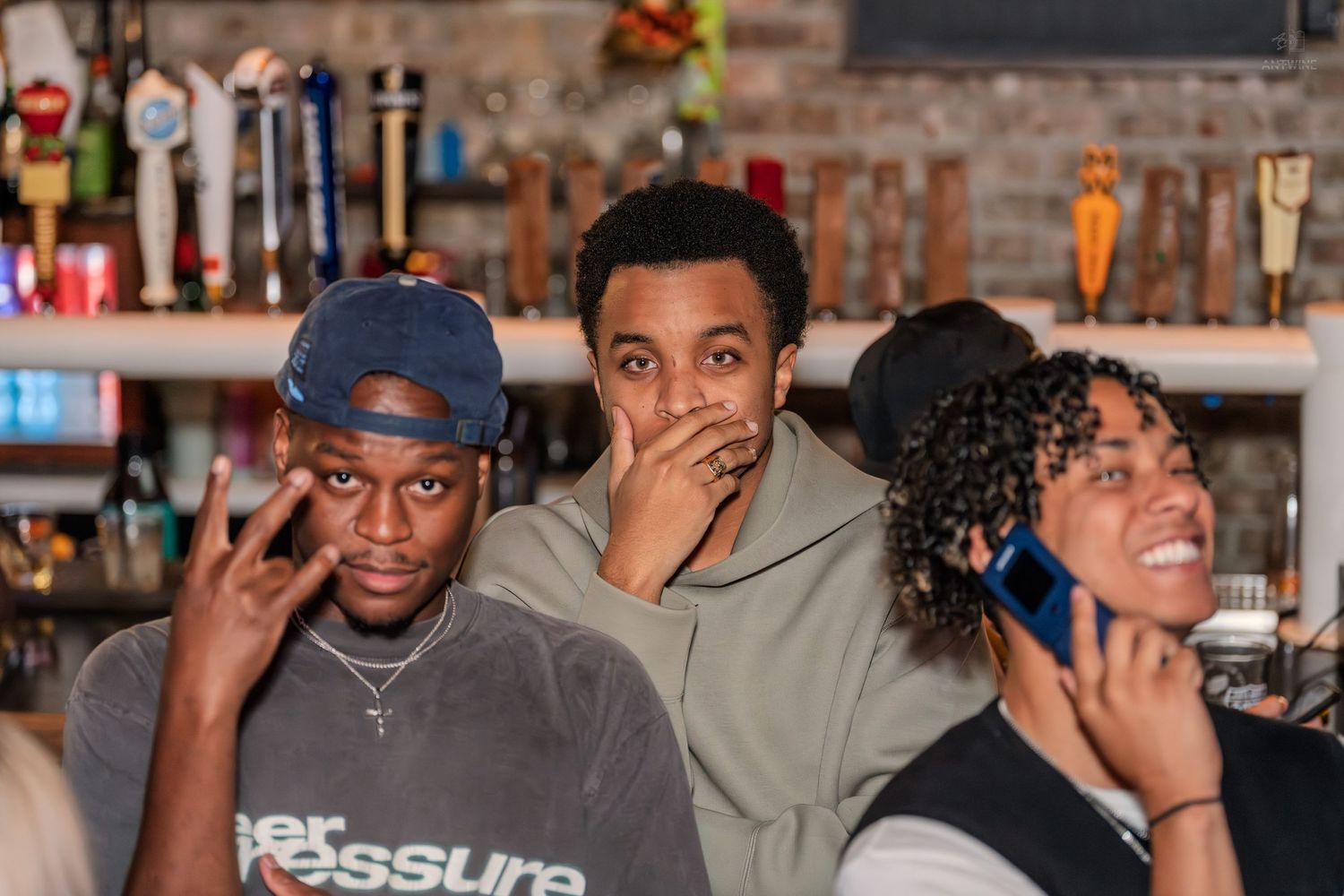 Three young friends at a bar, one flashing peace sign, another covering mouth in surprise, third smiling.