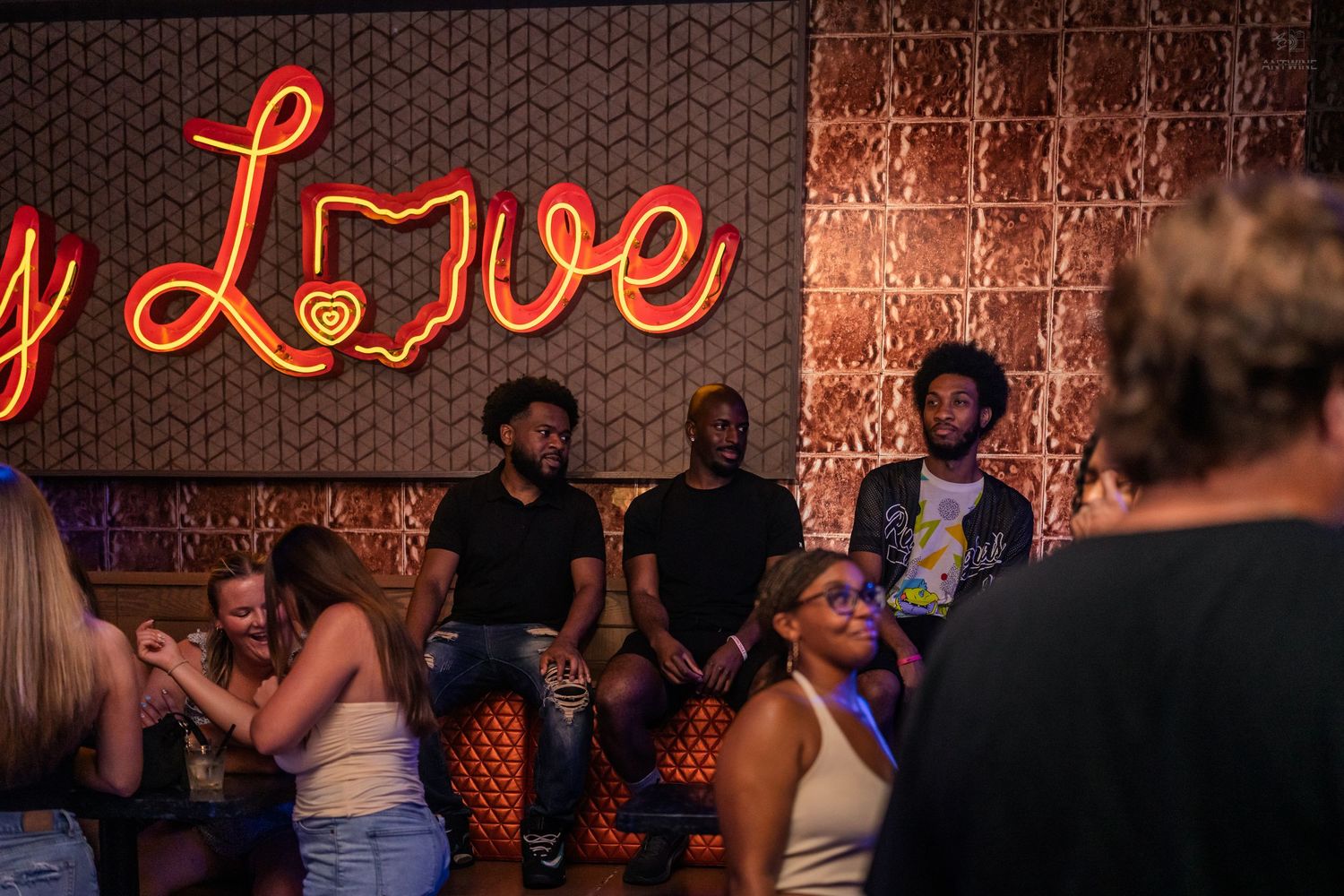 Friends gathered beneath a neon Love sign in a stylish nightclub lounge.