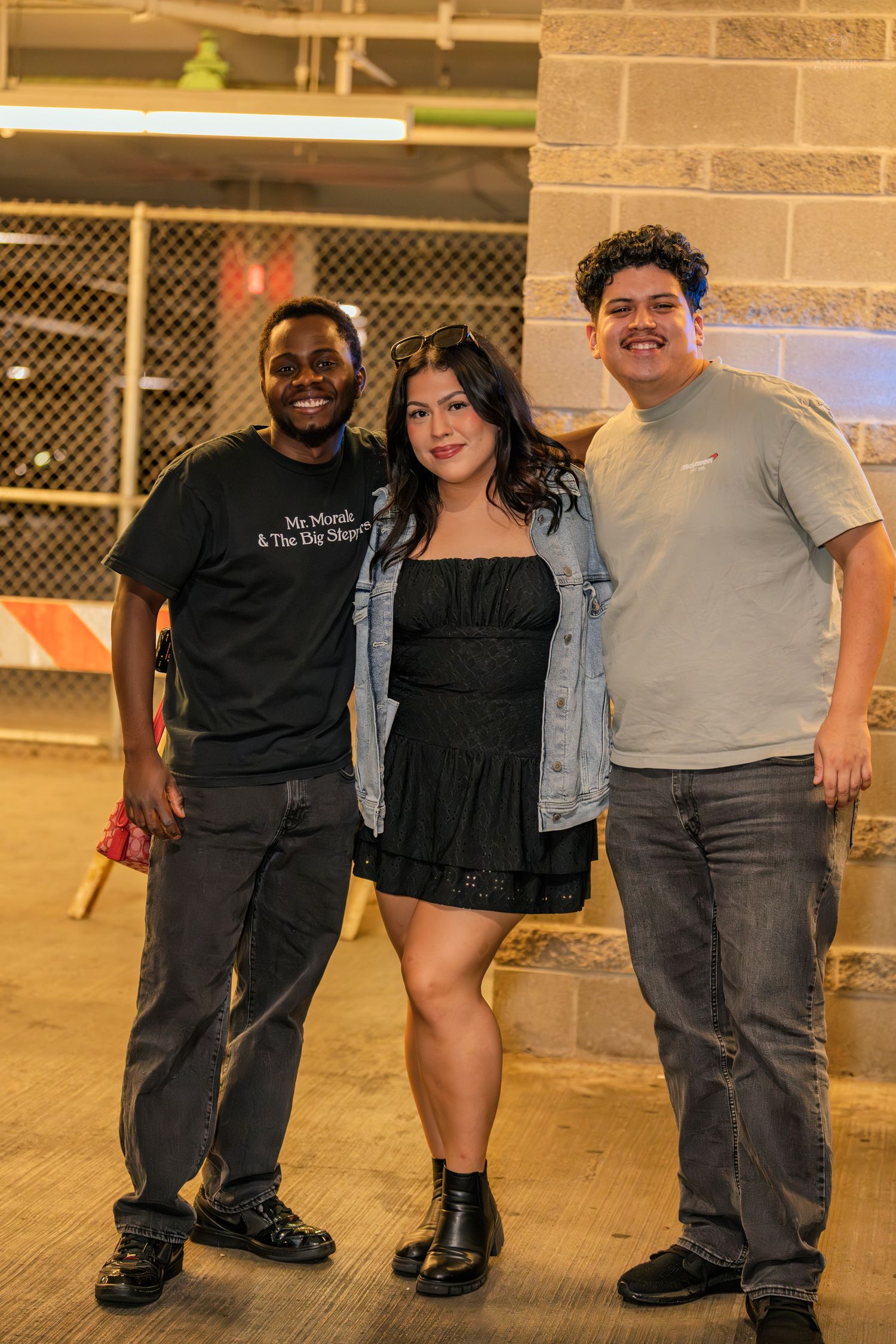 Three friends smiling and posing together in a parking garage at a nighttime event.