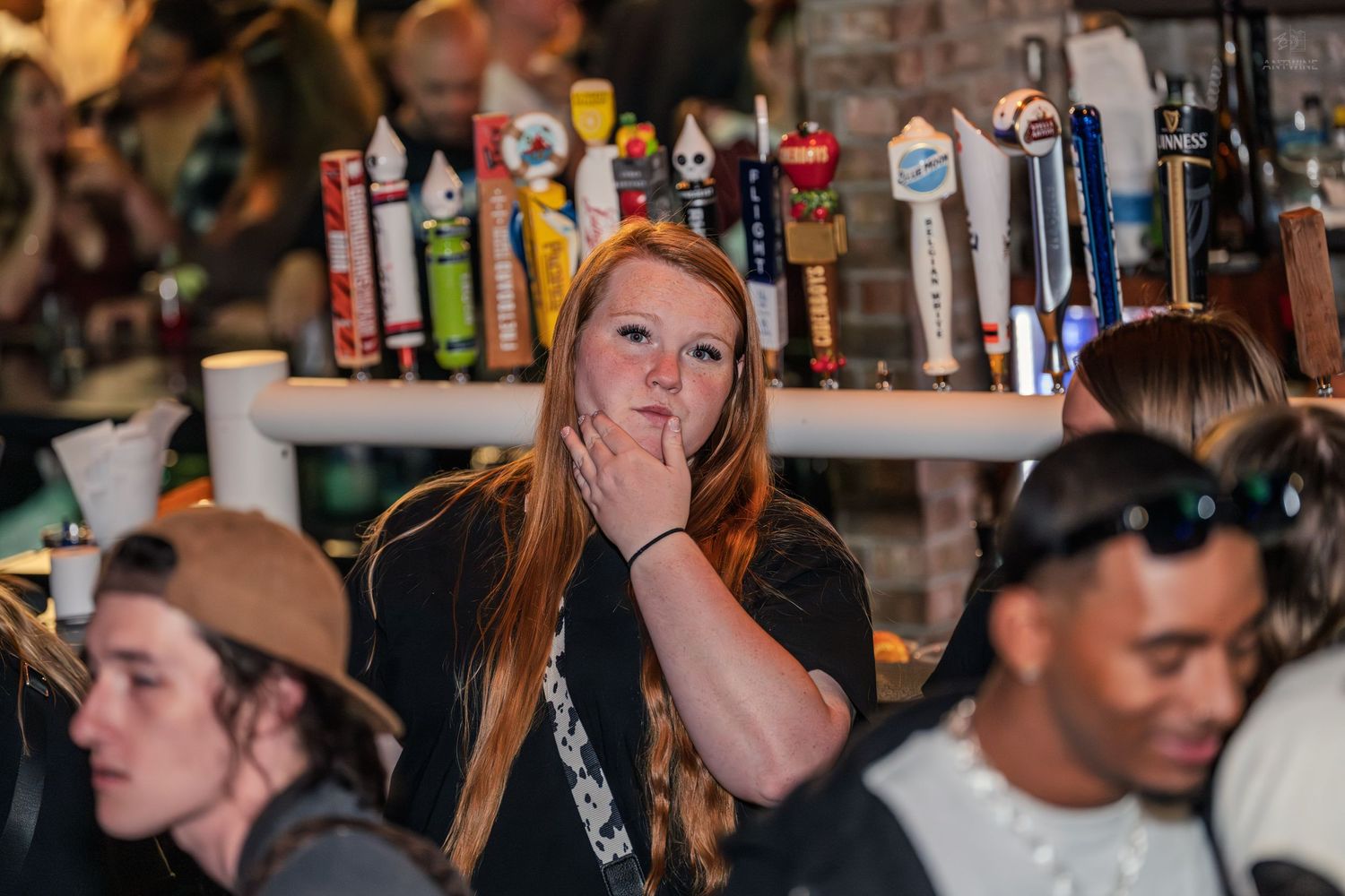 Redheaded woman covering her mouth in surprise near a row of draft beer taps at a busy bar.