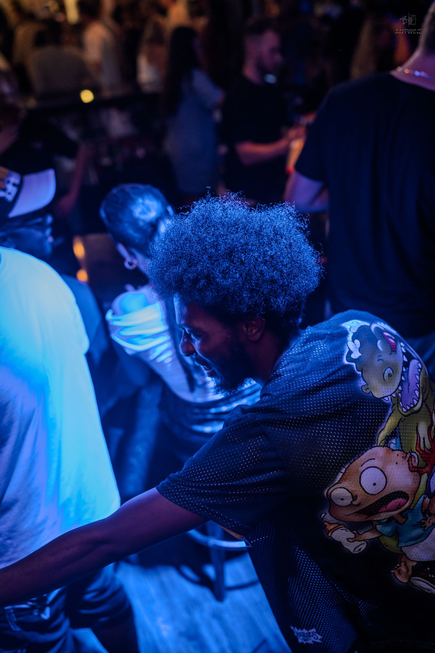 Man with blue-tinted afro wearing a graphic tee dancing at a nightclub event.