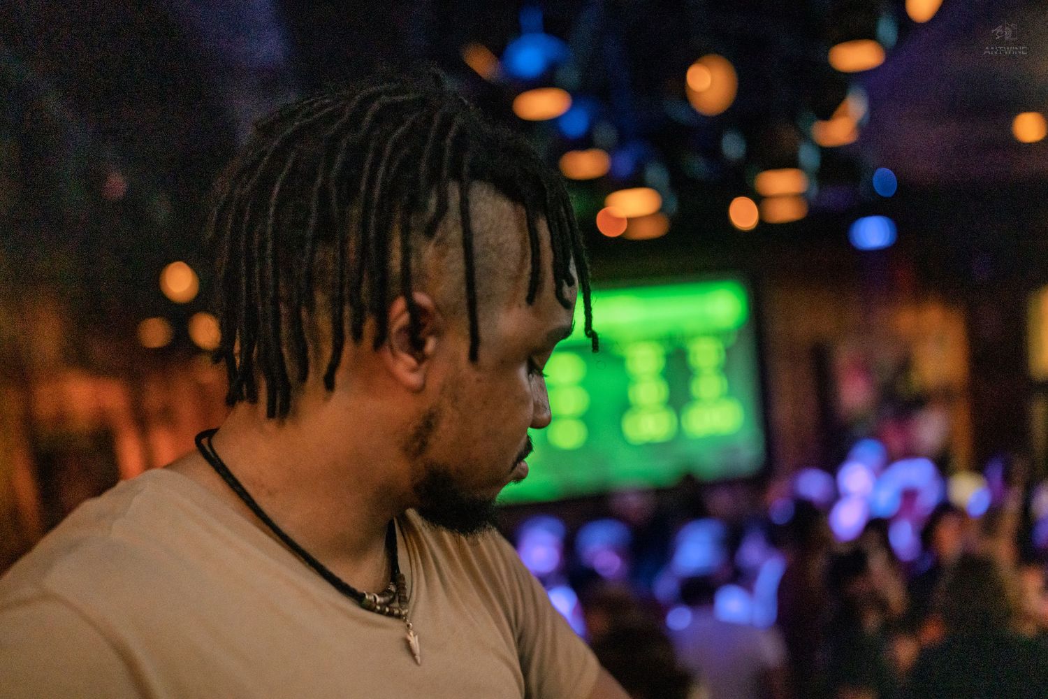 A man with dreadlocks looks down in a dark nightclub with green scoreboard lights in the background.