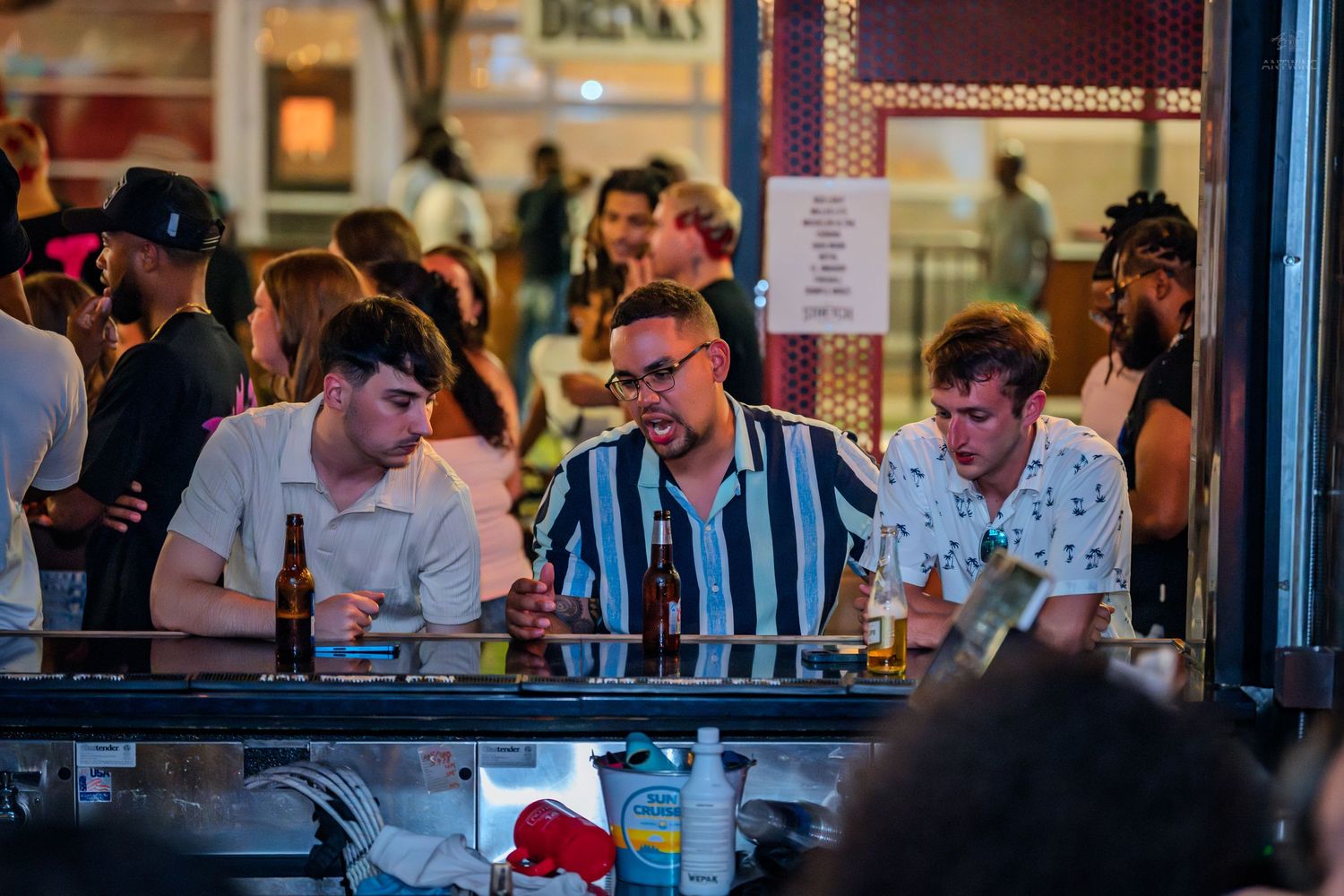 Three men socializing at a busy bar counter with beer bottles at night.