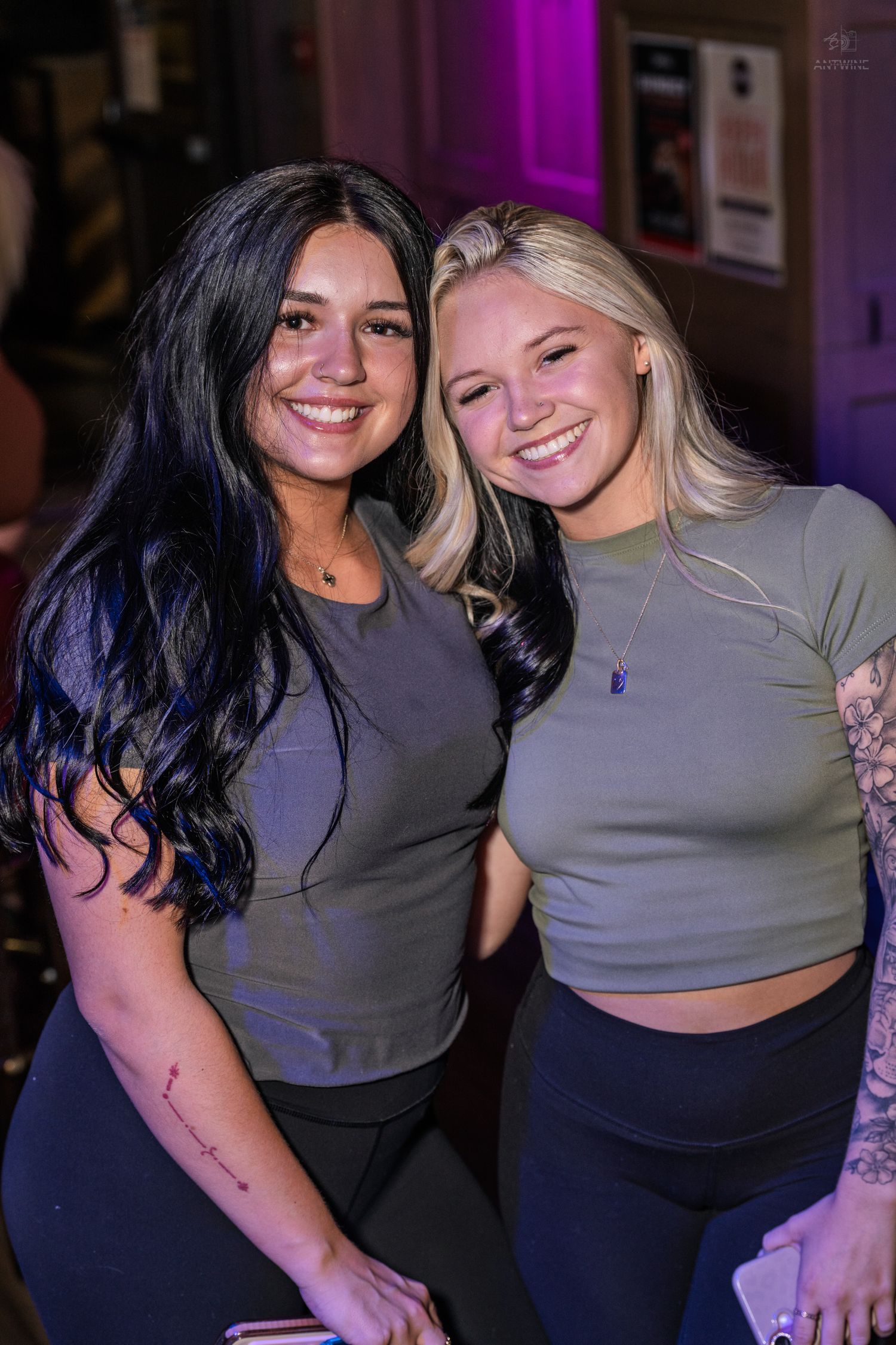 Two young women smile together at a bar, wearing matching olive green and gray outfits with black pants.