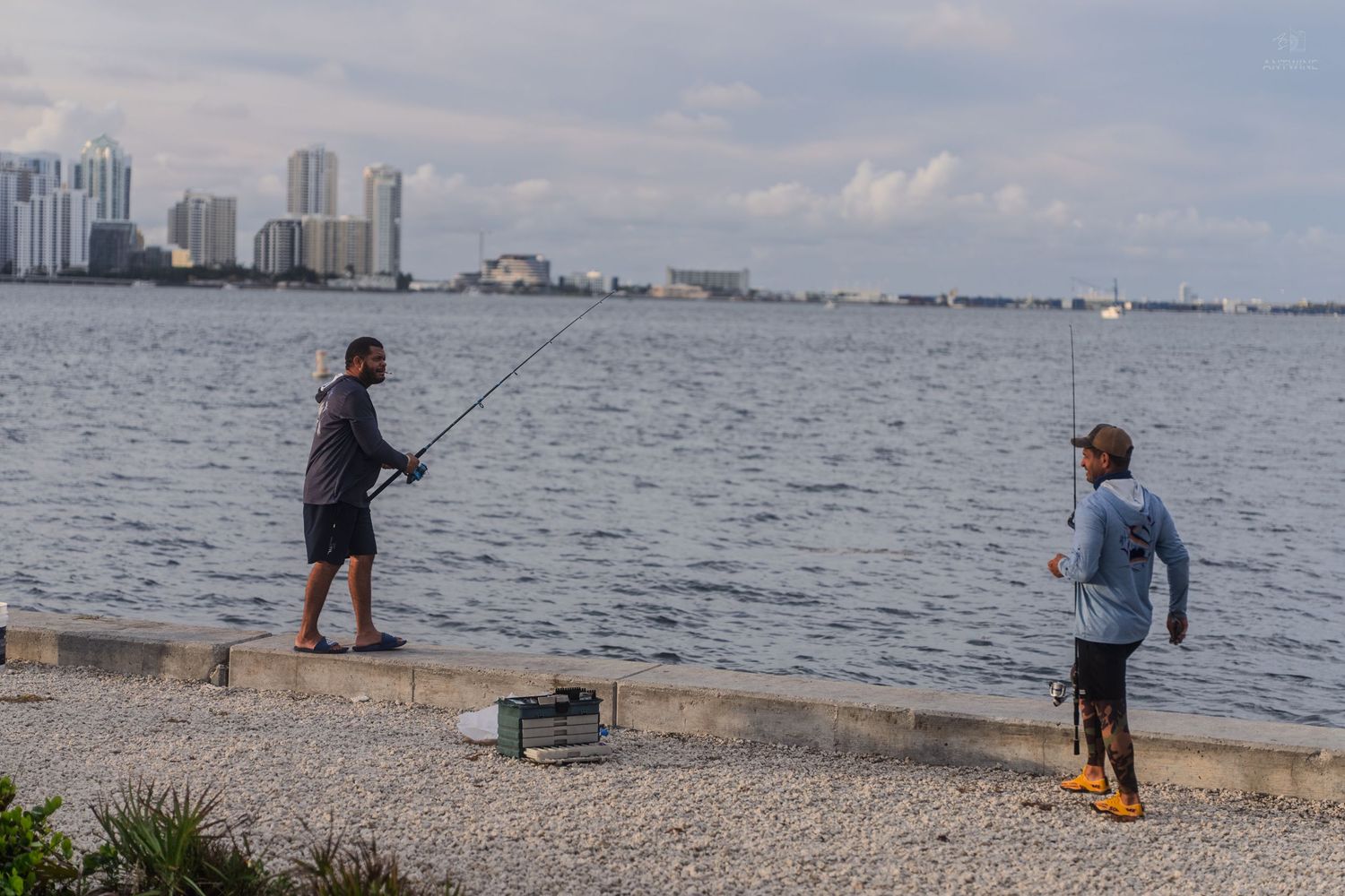 Two fishermen casting lines from rocky seawall with Miami downtown skyline across Biscayne Bay.