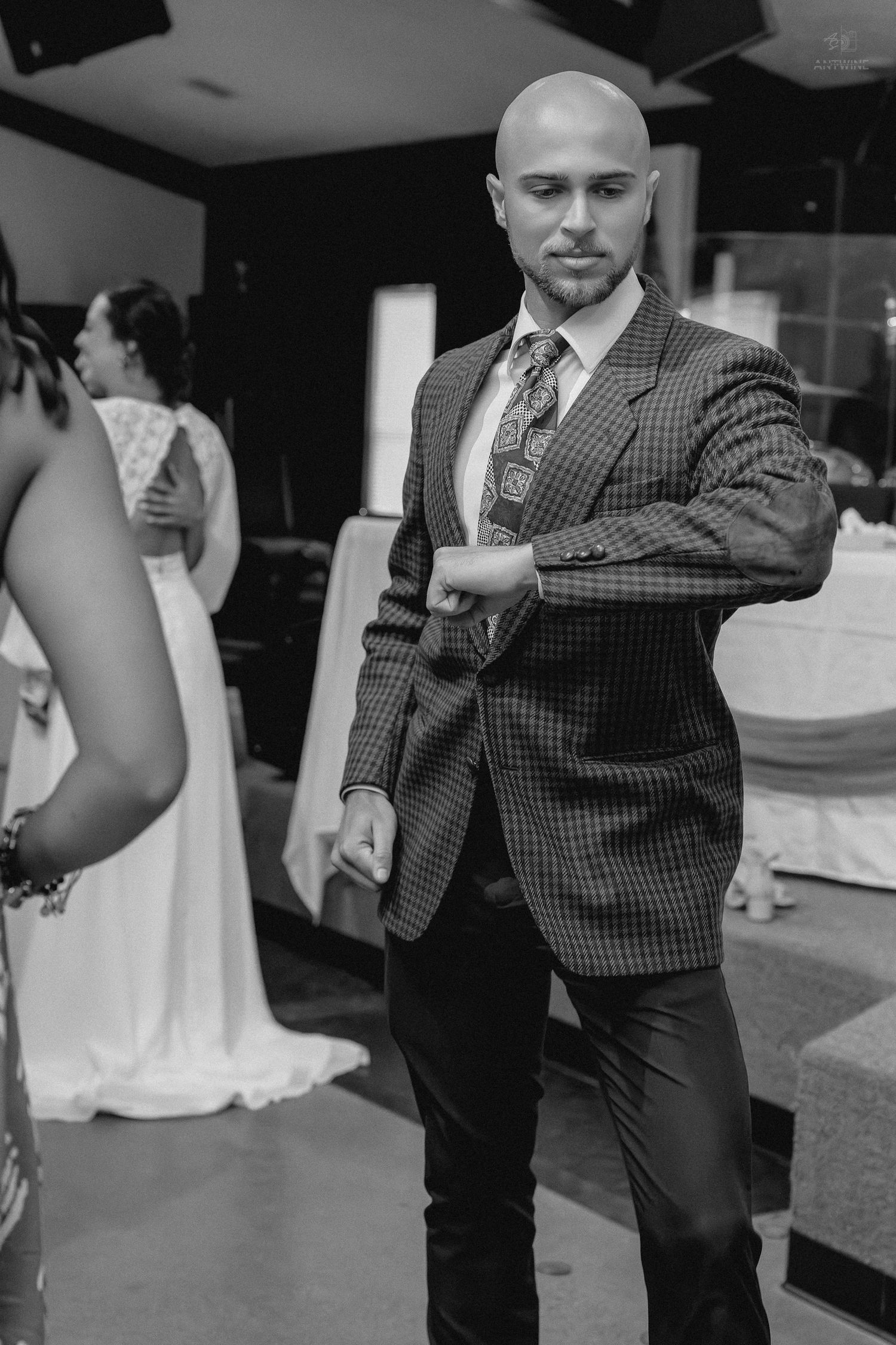 Black and white photo of bald man in checkered blazer dancing at formal wedding reception.
