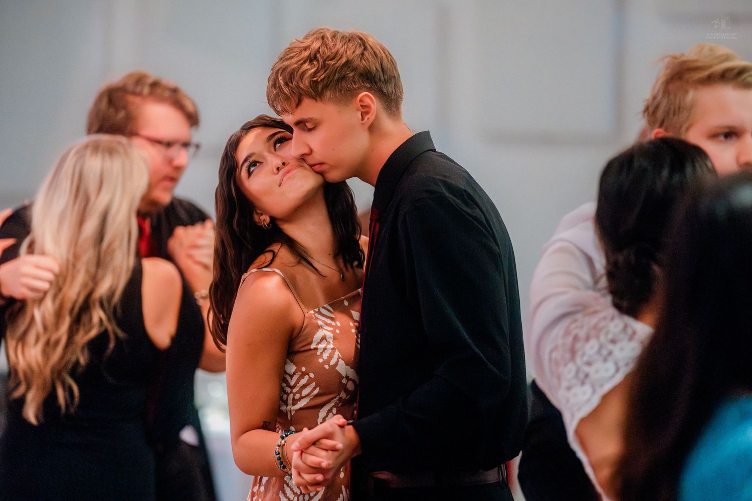 A young couple shares a romantic kiss while dancing at a formal event surrounded by other guests.