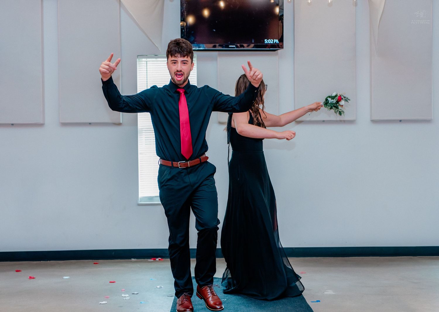 A couple dances at a formal event, man in black suit with red tie giving thumbs up, woman in black gown twirling.