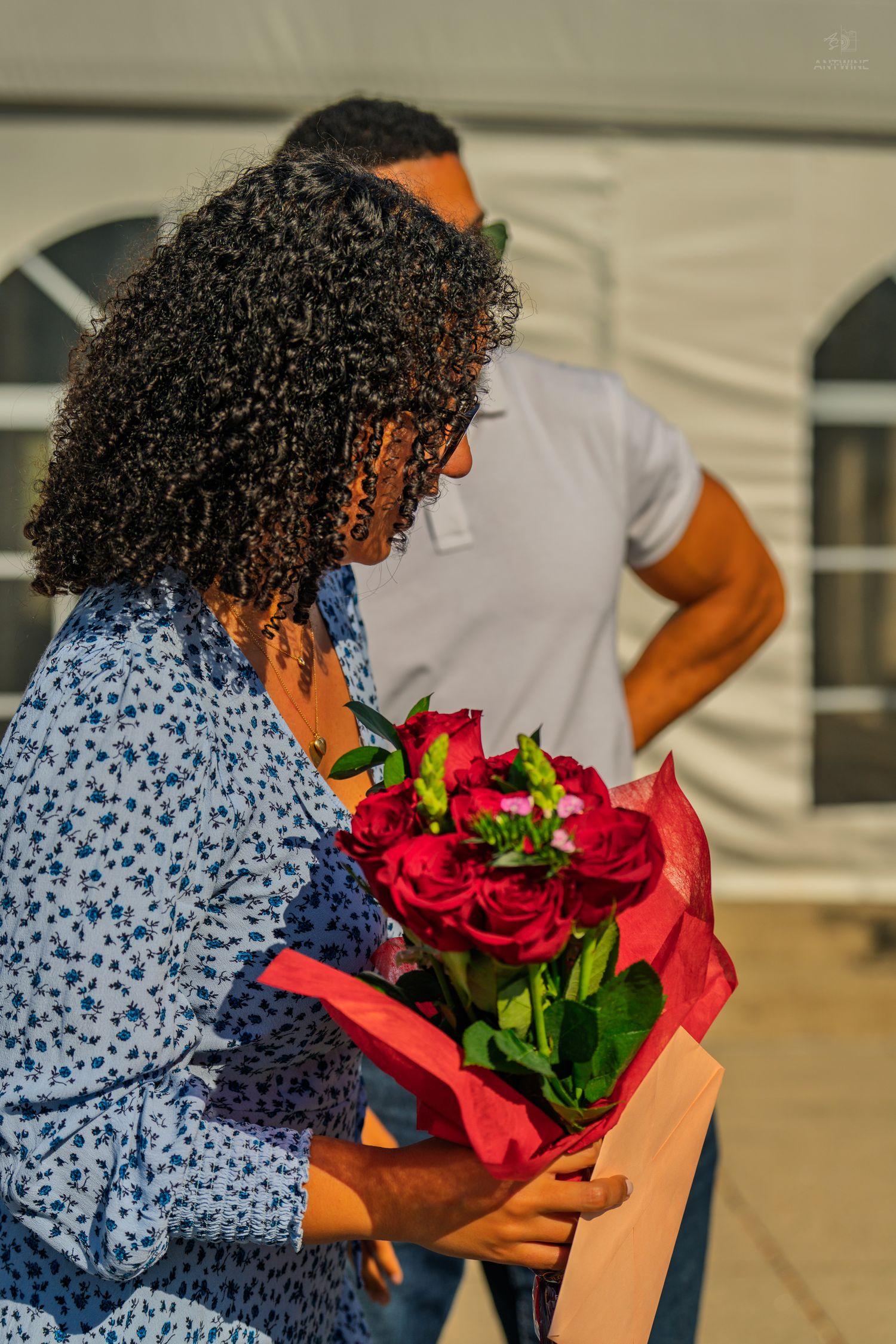 A woman with curly hair holds a bouquet of red roses while a man in a white polo stands behind her.