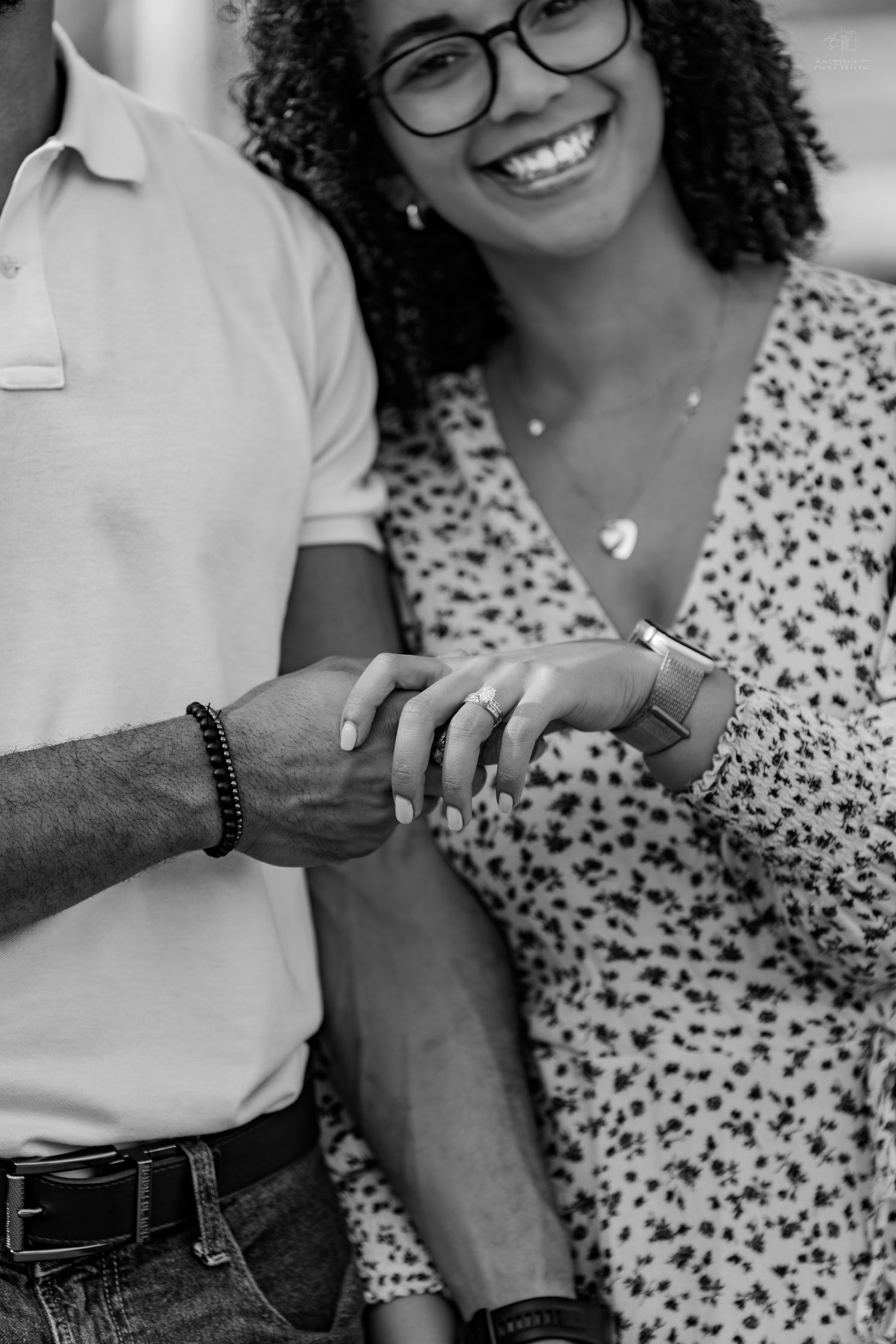 Black and white close-up of a joyful woman in glasses displaying an engagement ring while holding her partners hand.