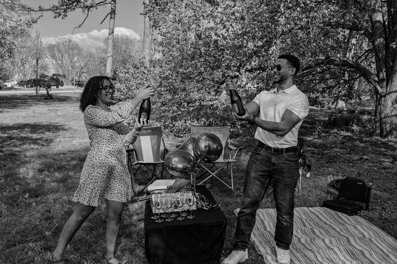 Black and white photo of a couple holding champagne bottles during an outdoor picnic celebration in a park.